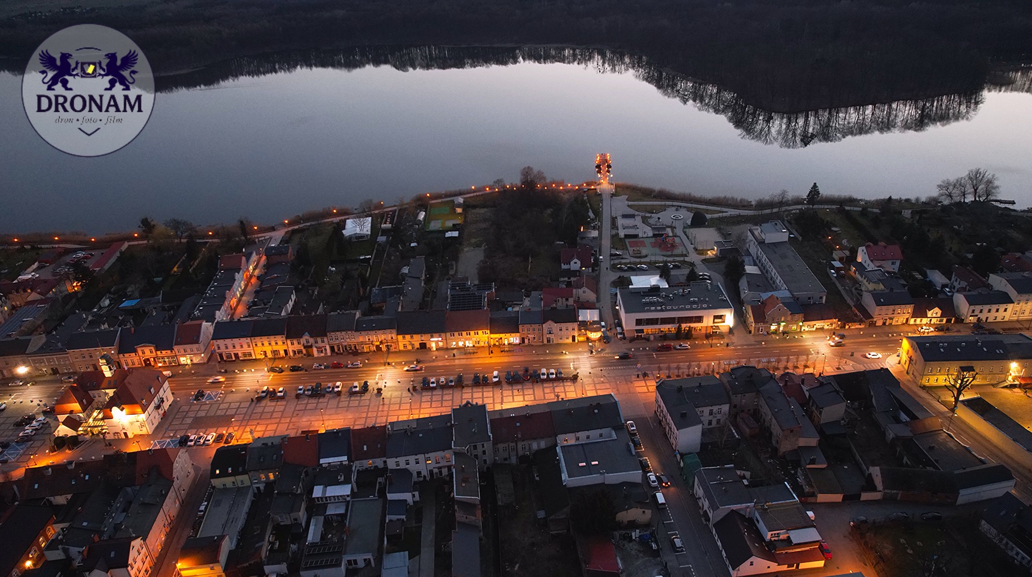 Nocny widok z drona na miasto nad jeziorem, z oświetlonymi budynkami i placem. W tle ciemne drzewa i woda. Logo firmy w lewym górnym rogu.