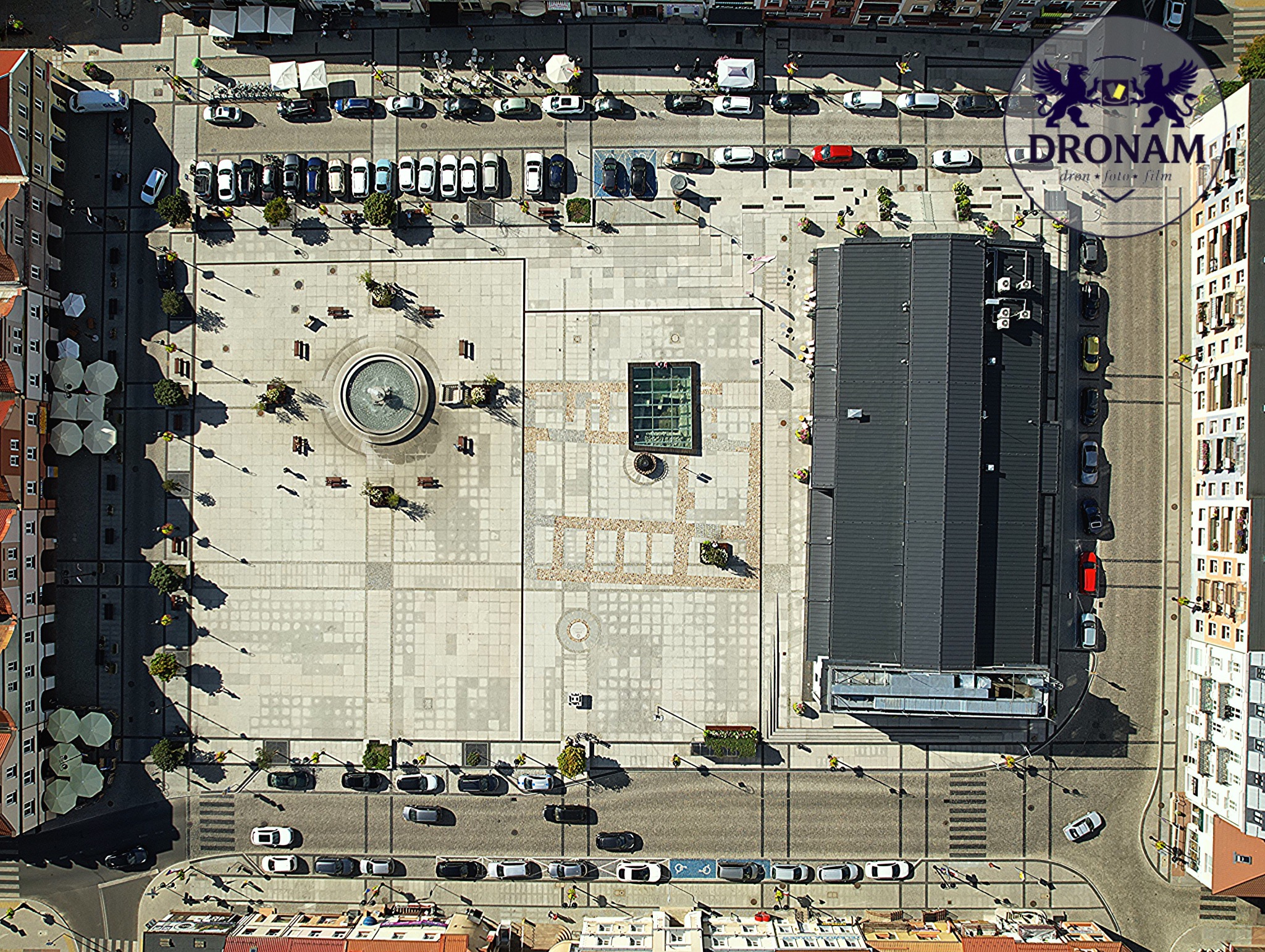 Widok z drona na rynek miejski z fontanną, budynkami i zaparkowanymi samochodami. Geometryczny układ placu, cień rzucany przez budynek. Logo firmy w prawym górnym rogu.