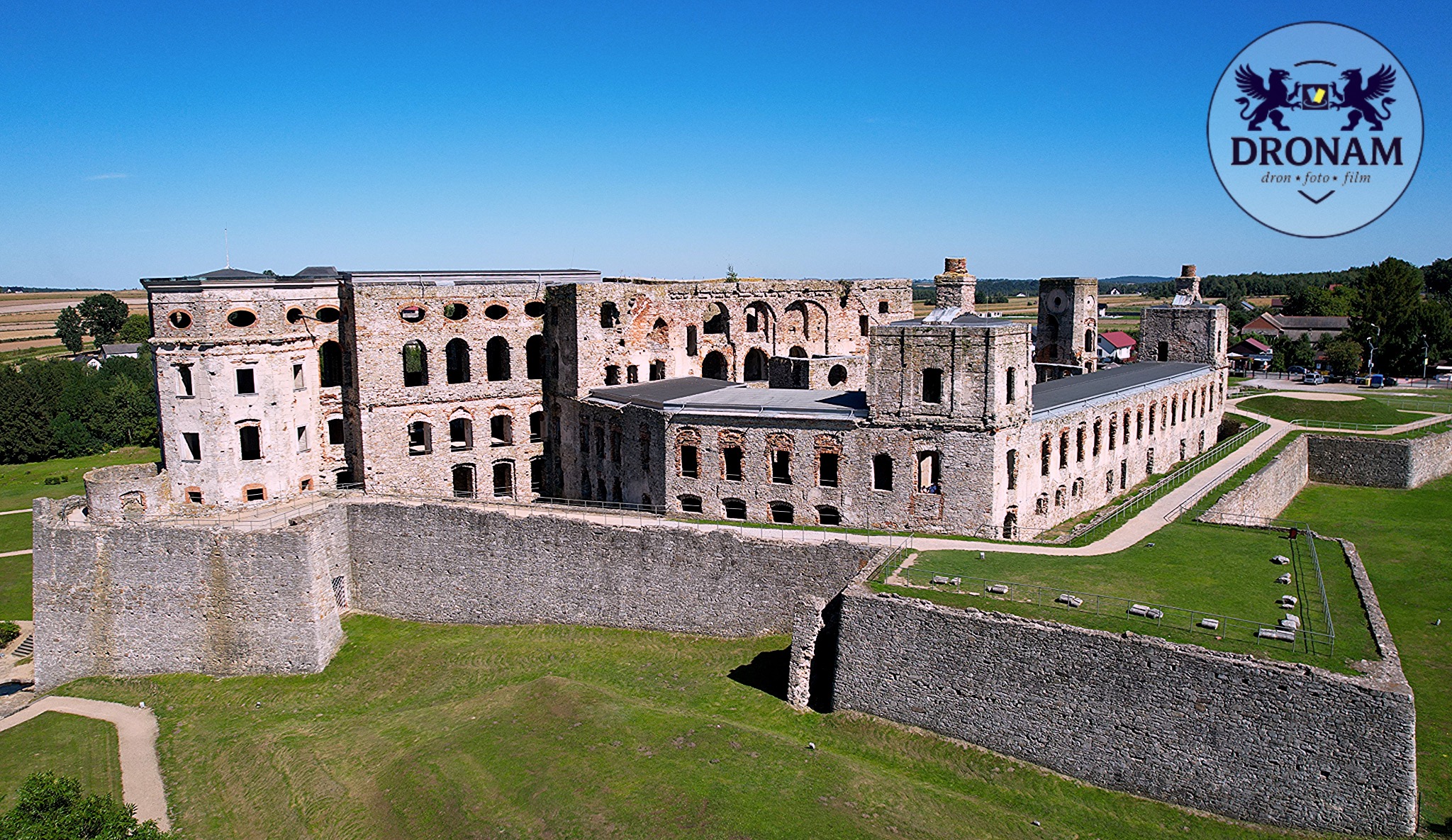 Panorama z drona na ruiny zamku Krzyżtopór w Ujeździe, z widocznym dziedzińcem i fragmentami murów obronnych. Logo firmy Dronam w prawym górnym rogu.