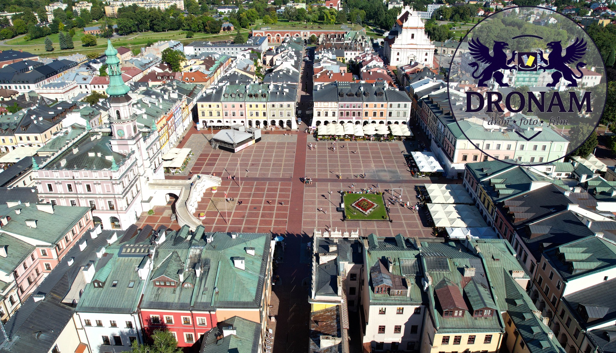 Panorama Zamościa z lotu ptaka, ukazująca Rynek Wielki z kolorowymi kamienicami, ratuszem i katedrą. Widoczny logotyp firmy Dronam.