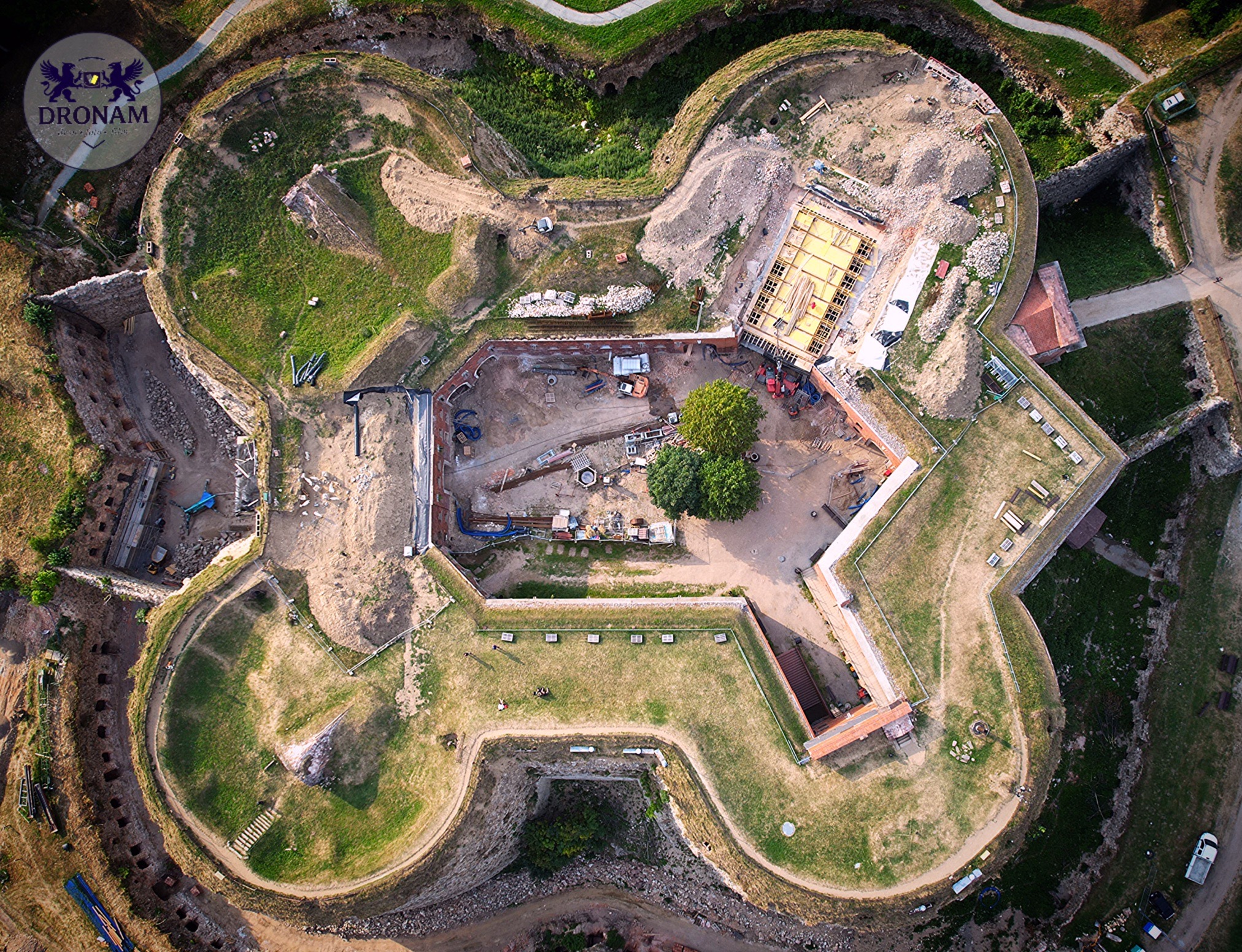 Fort w trakcie renowacji, widok z drona. Zewnętrzne wały ziemne porośnięte trawą, wewnątrz plac budowy z maszynami i materiałami. Unikalny kształt gwiazdy.