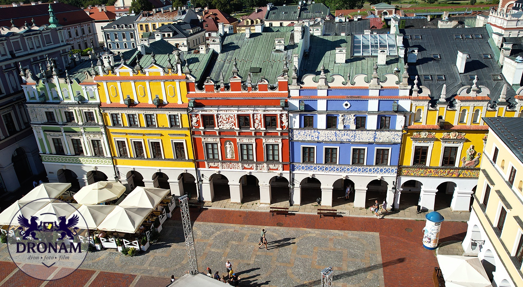 Kolorowe kamienice z podcieniami na Rynku Wielkim w Zamościu, widok z góry. Czerwona cegła placu, białe parasole restauracji. Ujęcie z drona.