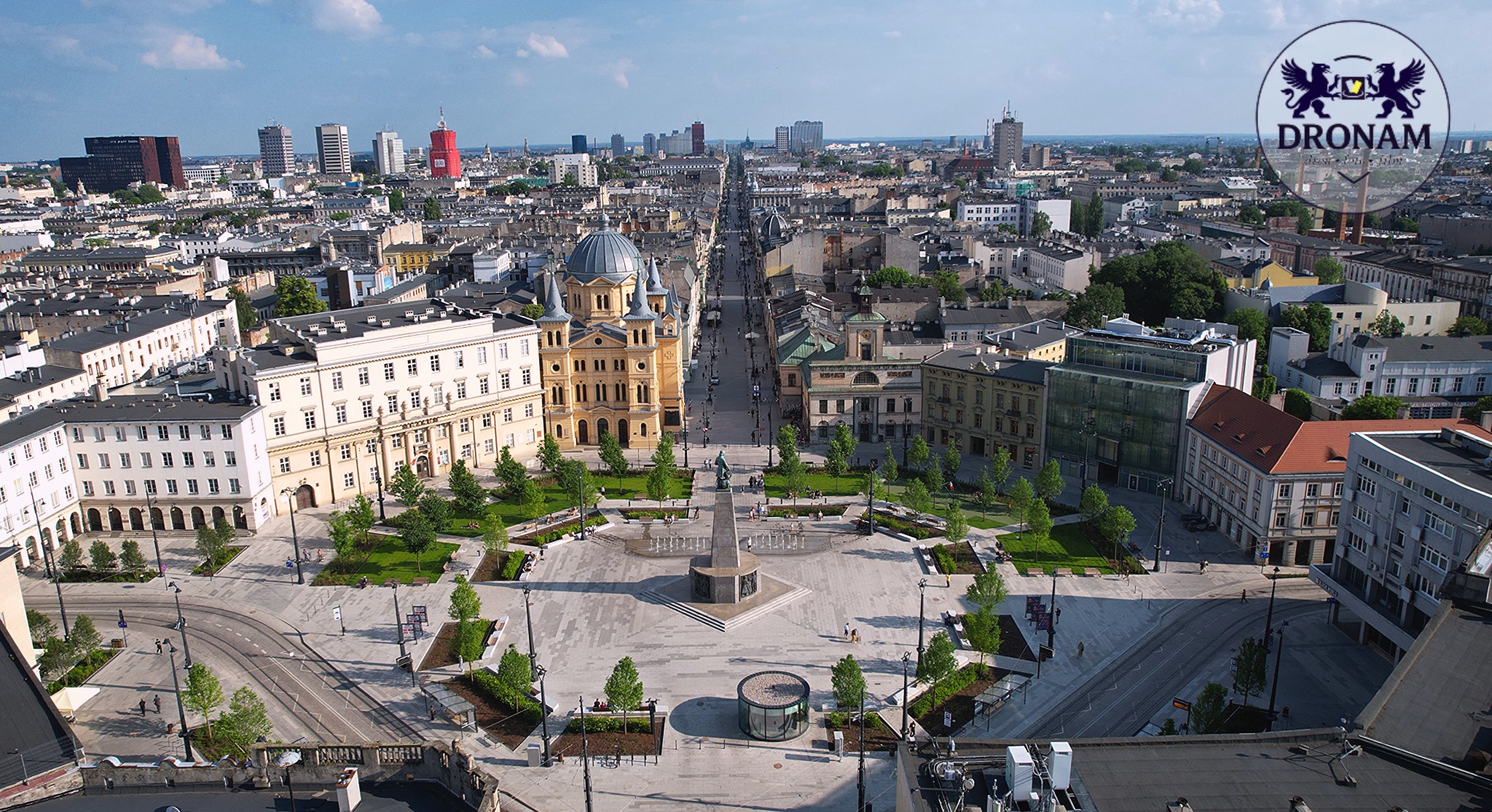Panorama miasta z lotu ptaka, centralny plac z fontanną i budynkami w stylu klasycystycznym, w tle nowoczesne wieżowce. Logo firmy w prawym górnym rogu.