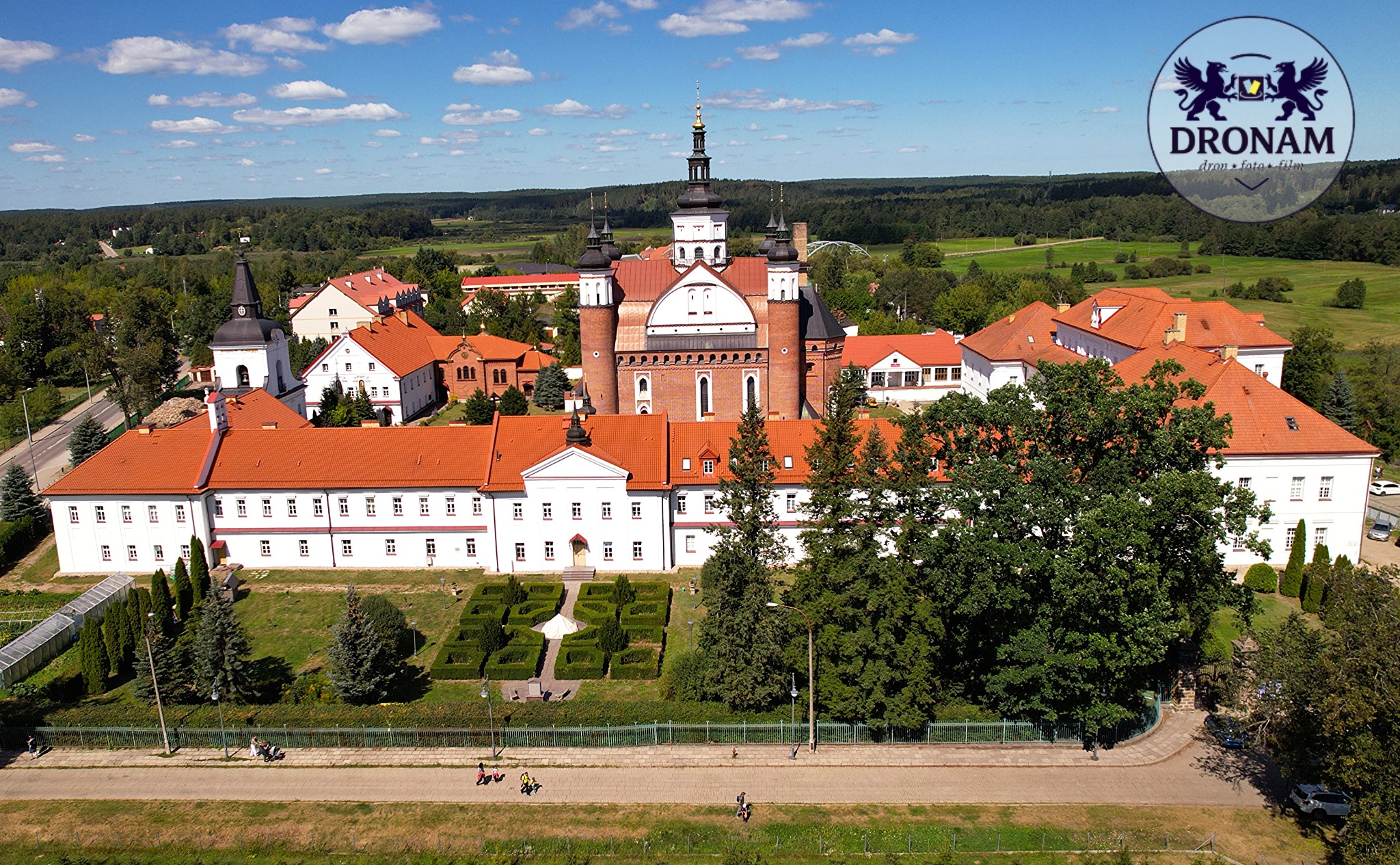 Szeroki kadr z drona na barokowy kompleks klasztorny z czerwonymi dachami, otoczony zielenią i parkiem. W prawym górnym rogu logo firmy Dronam.