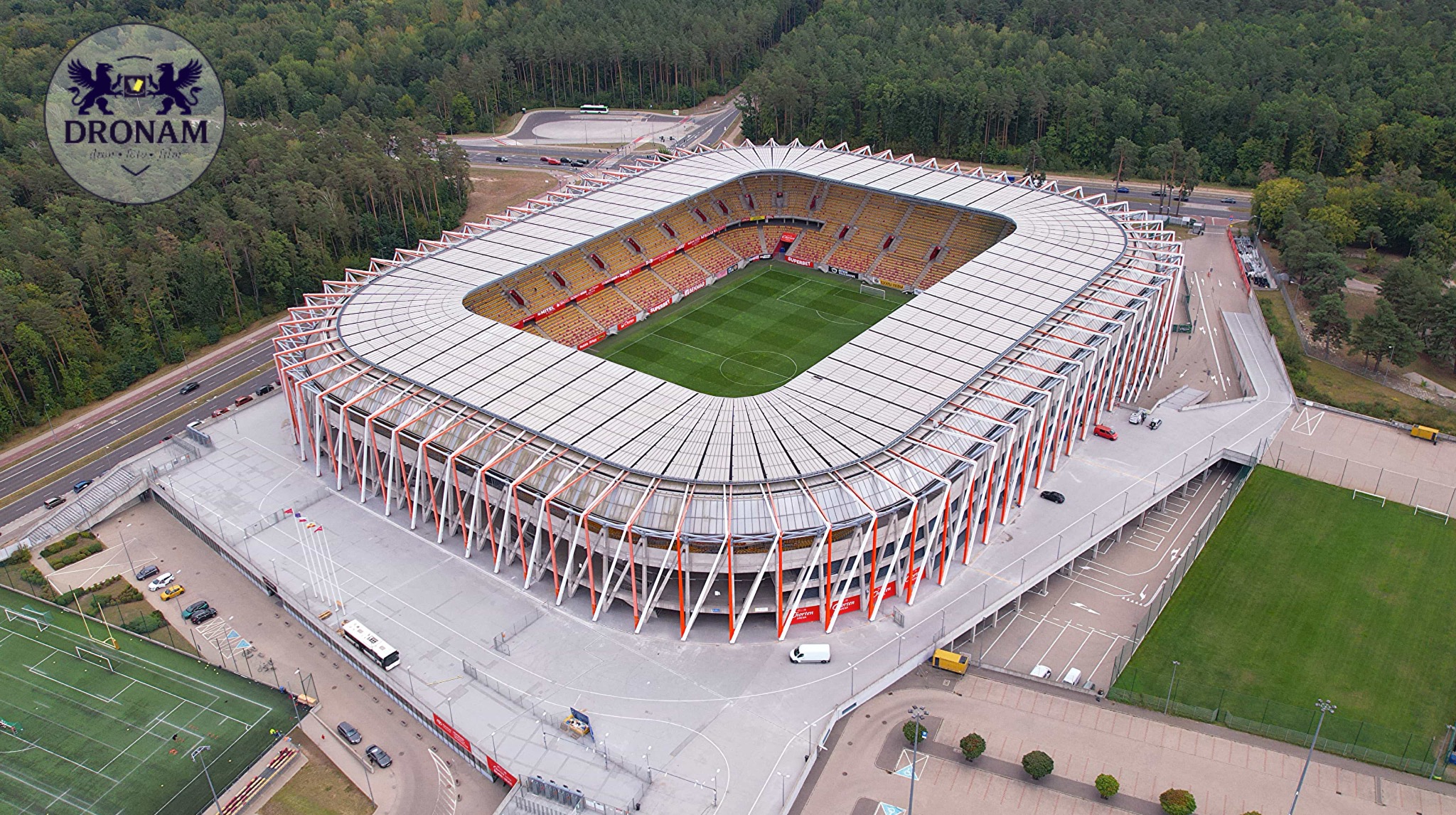Zdjęcie z drona przedstawia stadion z zieloną murawą i pomarańczowo-białą konstrukcją dachu. W tle widoczny las i droga. Ujęcie z góry, w słoneczny dzień.