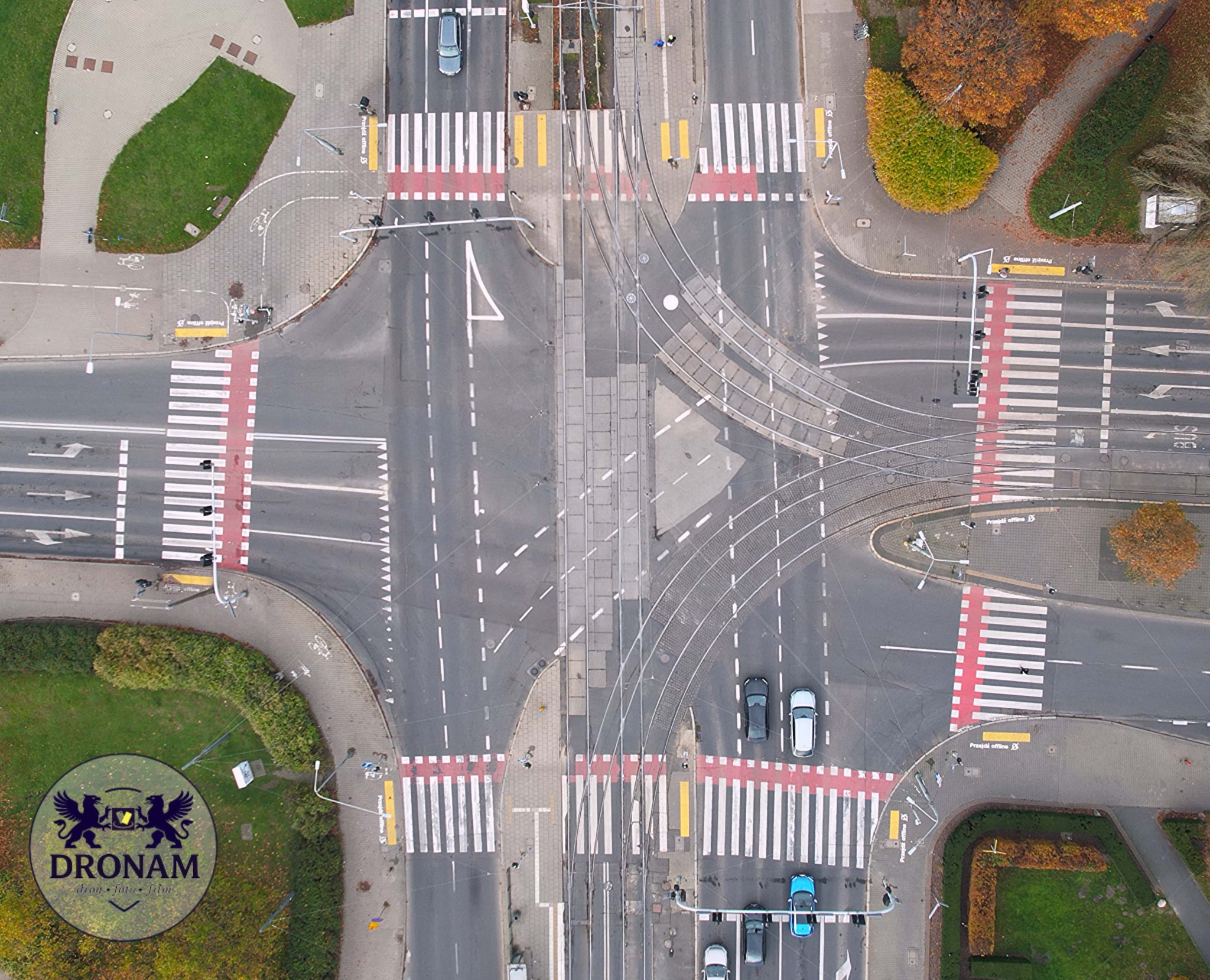Zdjęcie z drona przedstawia skrzyżowanie z torami tramwajowymi, przejściami dla pieszych i jezdnią. Widoczne samochody i zieleń. W lewym dolnym rogu logo firmy.