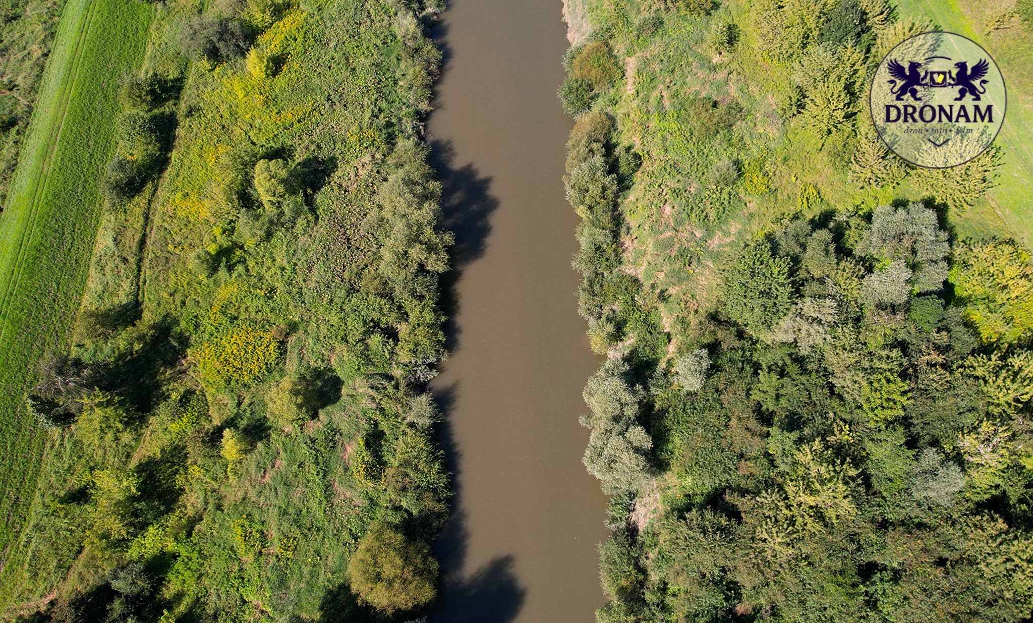 Zdjęcie z drona przedstawia rzekę płynącą przez zielony krajobraz. Widok z góry ukazuje koryto rzeki otoczone bujną roślinnością i polami. W prawym górnym rogu widoczne logo firmy.