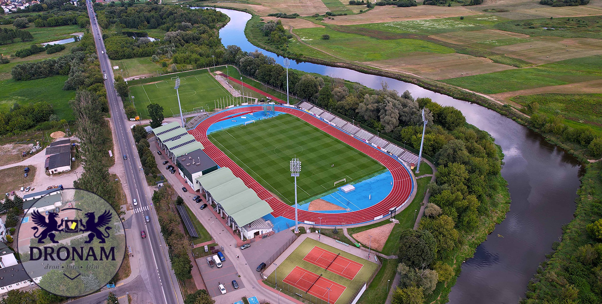 Widok z drona na stadion lekkoatletyczny z bieżnią, boiskiem, trybunami i kortami tenisowymi, położony nad rzeką, otoczony zielenią. W tle droga i zabudowania.