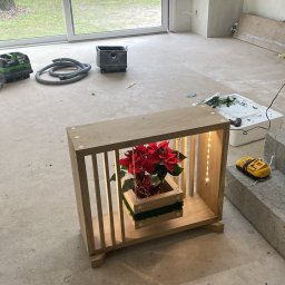 Kamil Głos - Drewniana konstrukcja z poinsecją w środku, wisząca na łańcuszkach, oświetlona lampkami, na tle surowego wnętrza w trakcie wykończenia. Widoczne narzędzia.