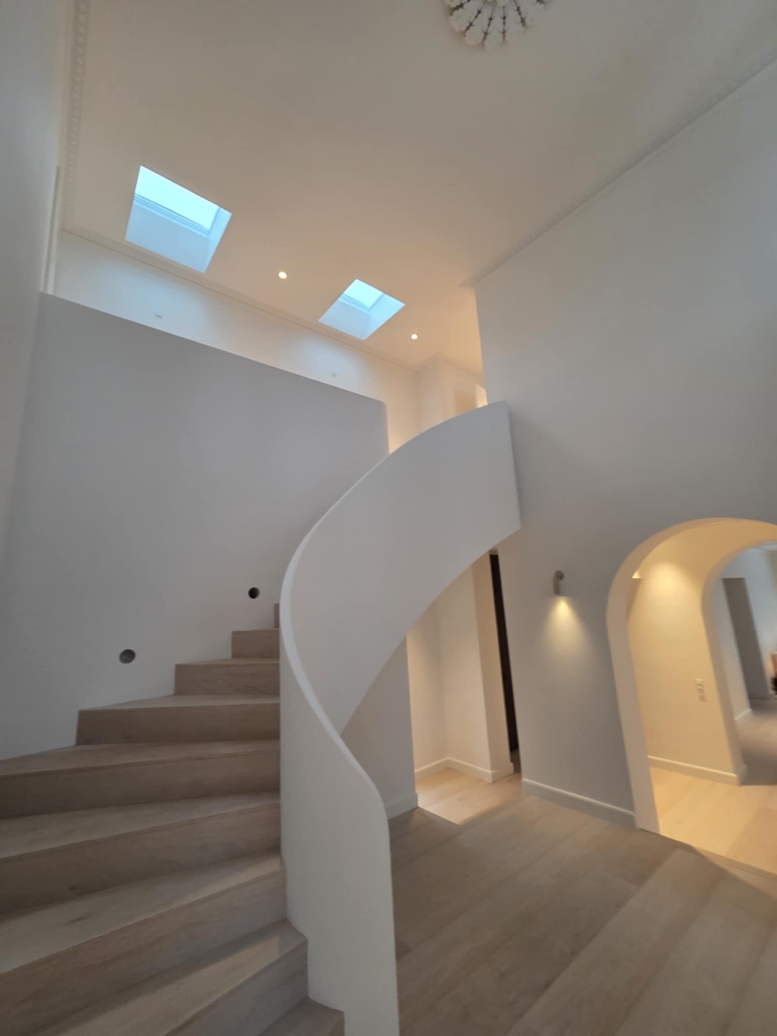Jasne drewniane schody spiralne w nowoczesnym, białym wnętrzu z oknami dachowymi i łukowym przejściem. Minimalistyczny design, ciepłe oświetlenie.