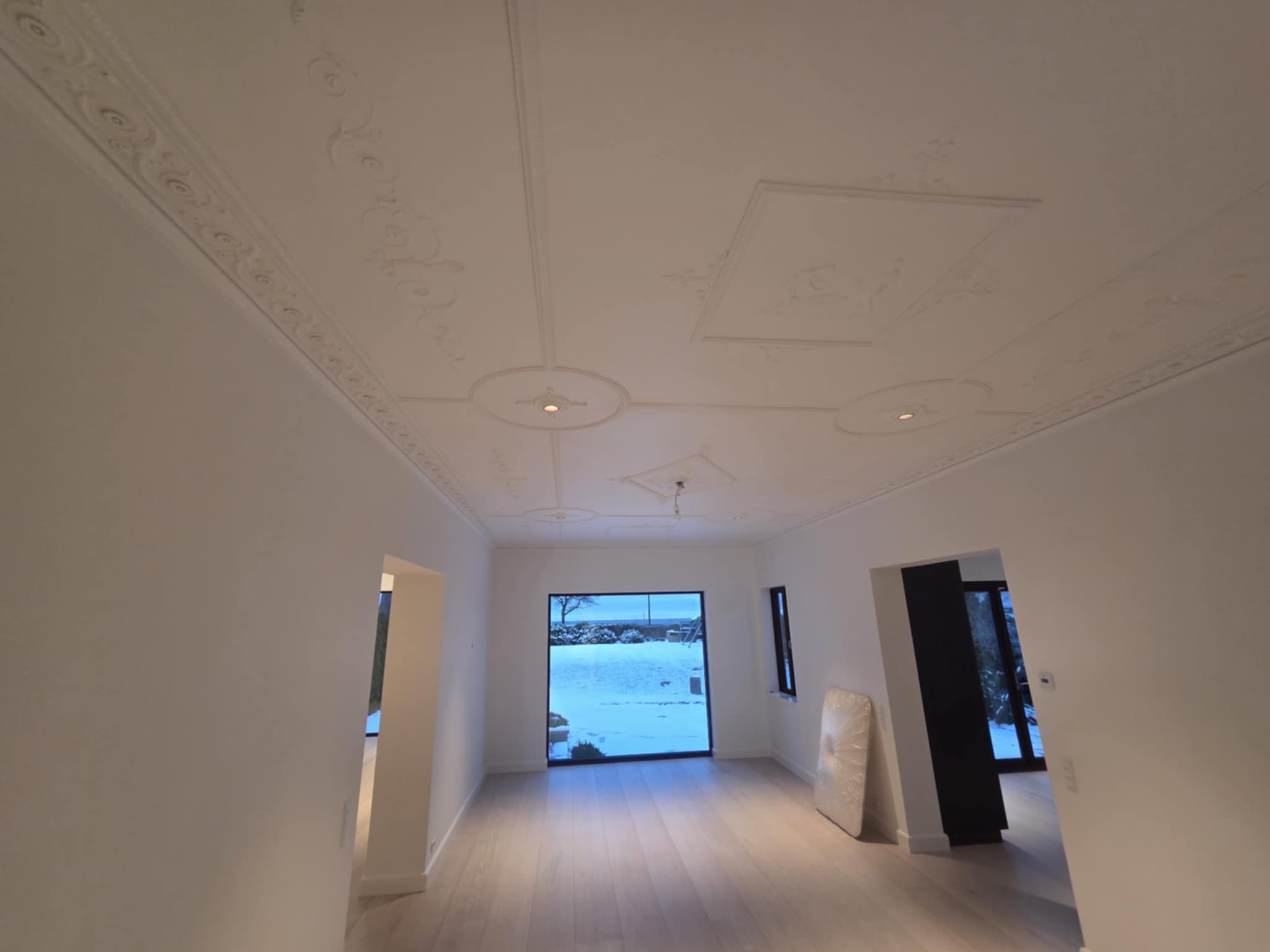 Białe, wykończone wnętrze z dekoracyjnym sufitem i oknem z widokiem na zimowy krajobraz. Parkiet na podłodze, minimalistyczny design.