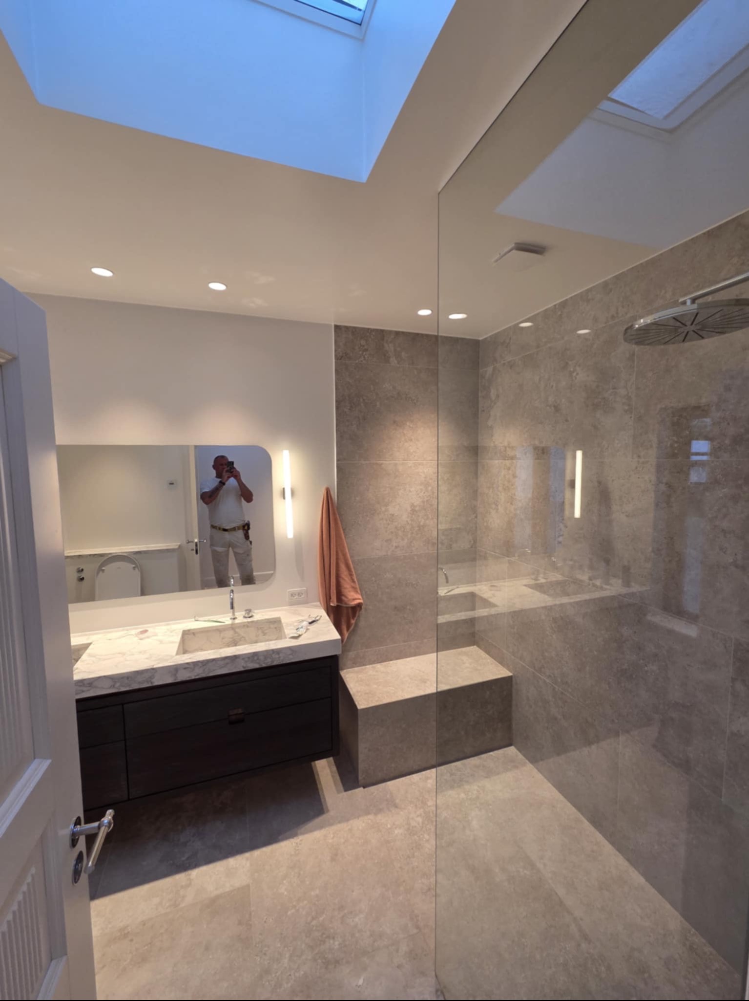 Nowoczesna łazienka z szarymi płytkami, kabiną prysznicową typu walk-in i marmurową umywalką. W lustrze odbija się osoba robiąca zdjęcie. Widoczne okno dachowe.