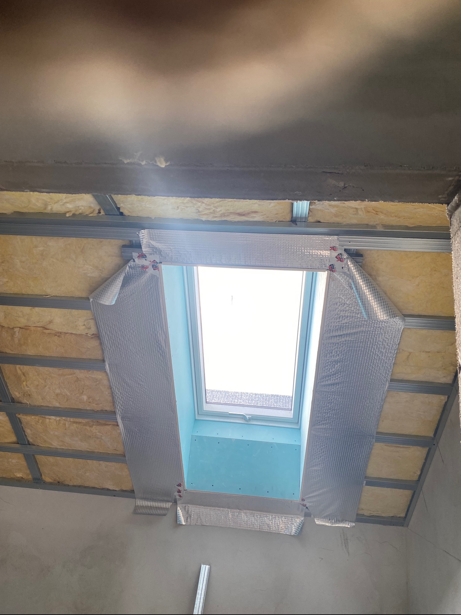 Montaż okna dachowego: izolacja wełną mineralną, folia aluminiowa, metalowy stelaż. Widok od dołu, surowy stan wykończenia ścian.