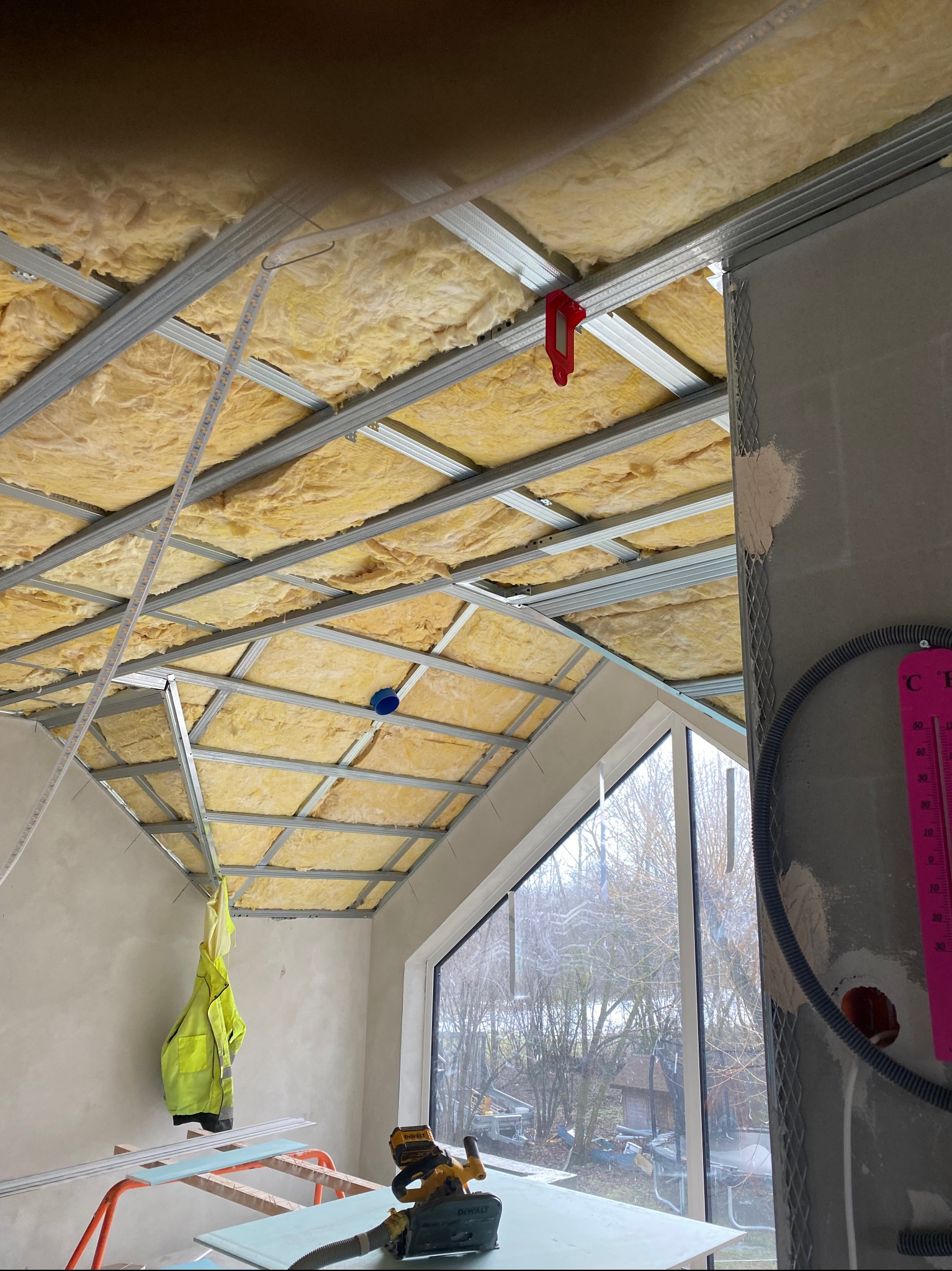 Sufit podwieszany z izolacją z wełny mineralnej, widoczne profile metalowe i narzędzia na tle okna. Prace wykończeniowe w trakcie realizacji.