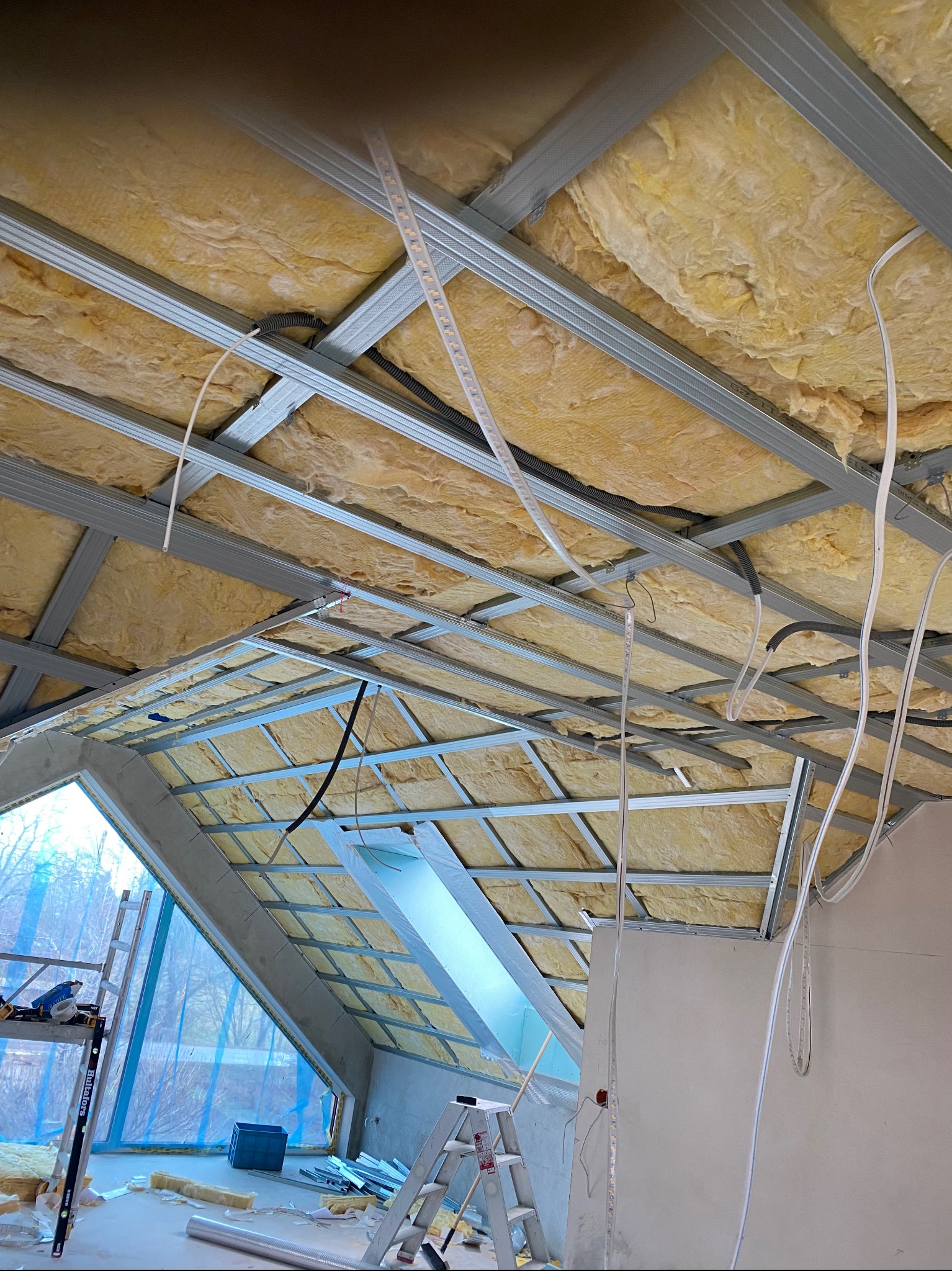 Skośny sufit poddasza w trakcie izolacji wełną mineralną, widoczne metalowe profile, okno dachowe i drabiny. Prace wykończeniowe w nowym domu.