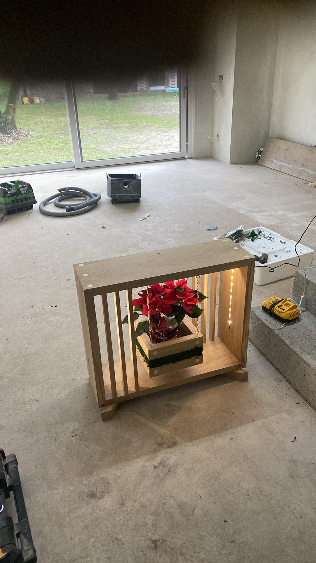 Drewniana konstrukcja z poinsecją w środku, wisząca na łańcuszkach, oświetlona lampkami, na tle surowego wnętrza w trakcie wykończenia. Widoczne narzędzia.