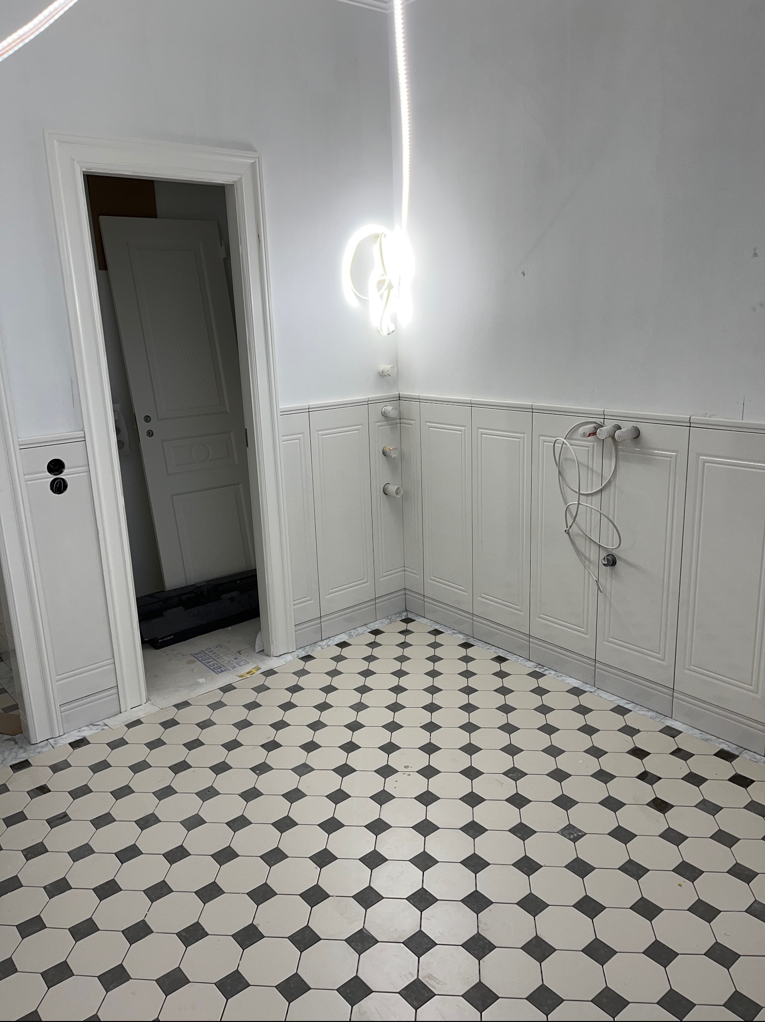 Wykończona łazienka z białą boazerią panelową i wzorzystą podłogą w stylu retro. Widoczne przygotowane punkty instalacji wodnej i elektrycznej.