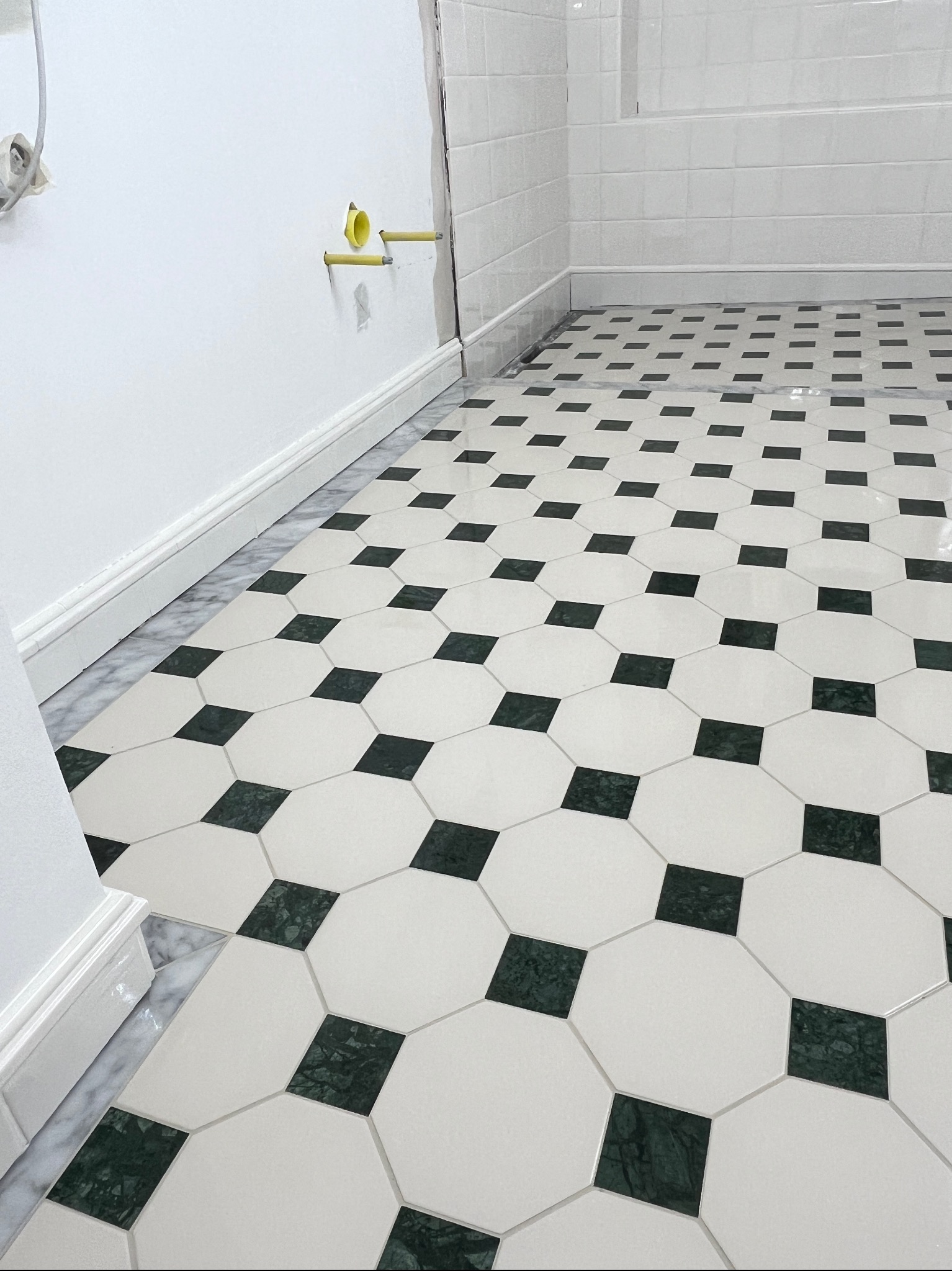 Ukończona podłoga w łazience z białych i zielonych płytek ułożonych w geometryczny wzór. Widoczne fragmenty ściany z instalacją hydrauliczną.