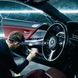 One Way Detailing - Wnętrze BMW: detailer w rękawicach czyści szczotką boczek drzwi. Widoczna kierownica, skórzane fotele i oświetlenie LED. Auto detailing w profesjonalnym studiu.
