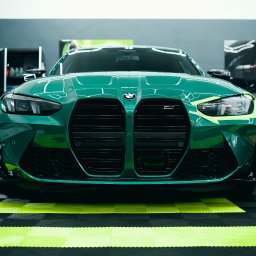 One Way Detailing - Zielony samochód BMW M4 G82 z czarnym grillem i żółtymi akcentami, przygotowywany do oklejania, widok z przodu na tle garażu z otwartymi drzwiami.