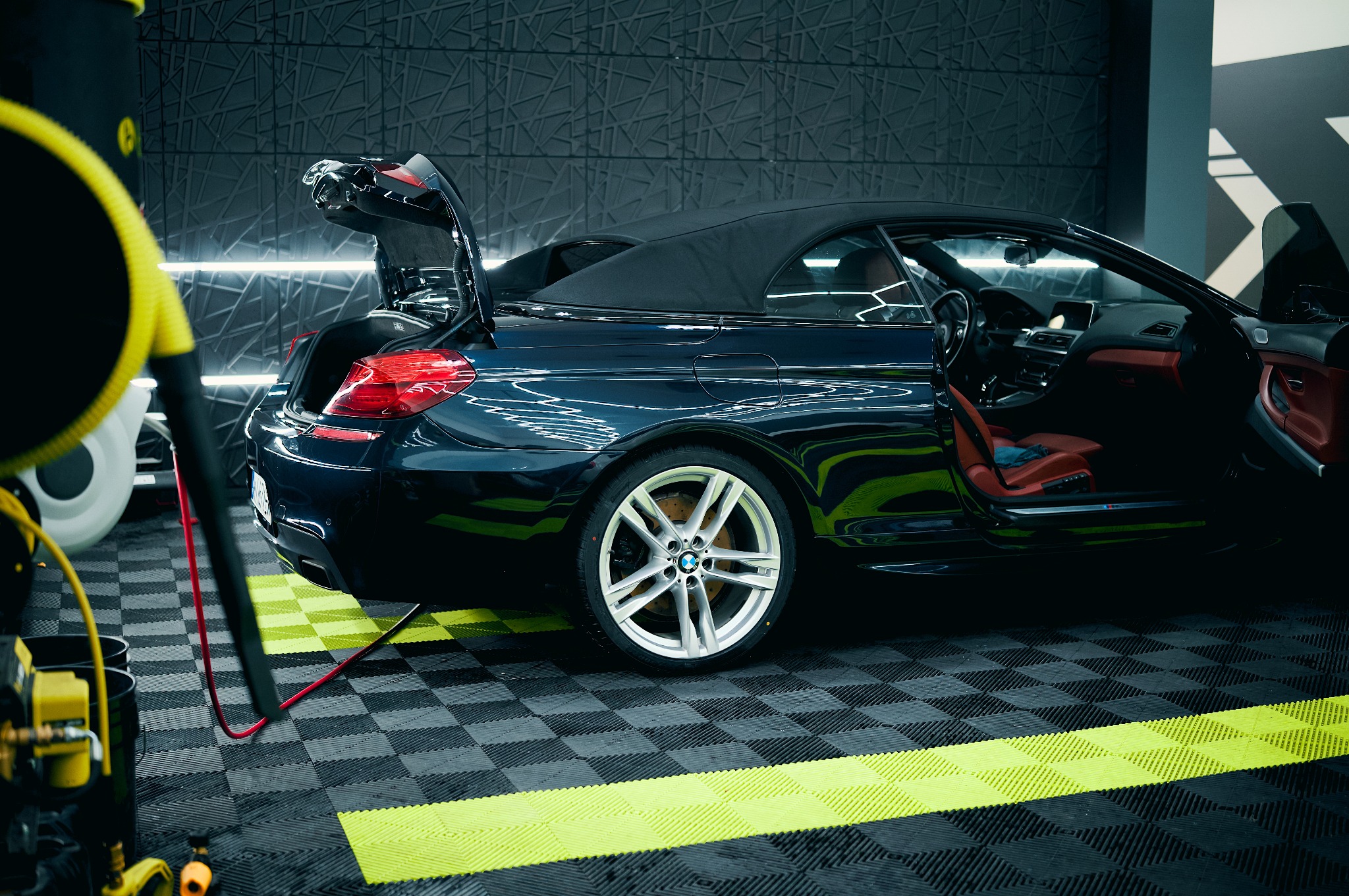 Błyszczący, granatowy kabriolet BMW z otwartym bagażnikiem i drzwiami, zaparkowany w nowoczesnej myjni z podłogą w szachownicę i oświetleniem LED.