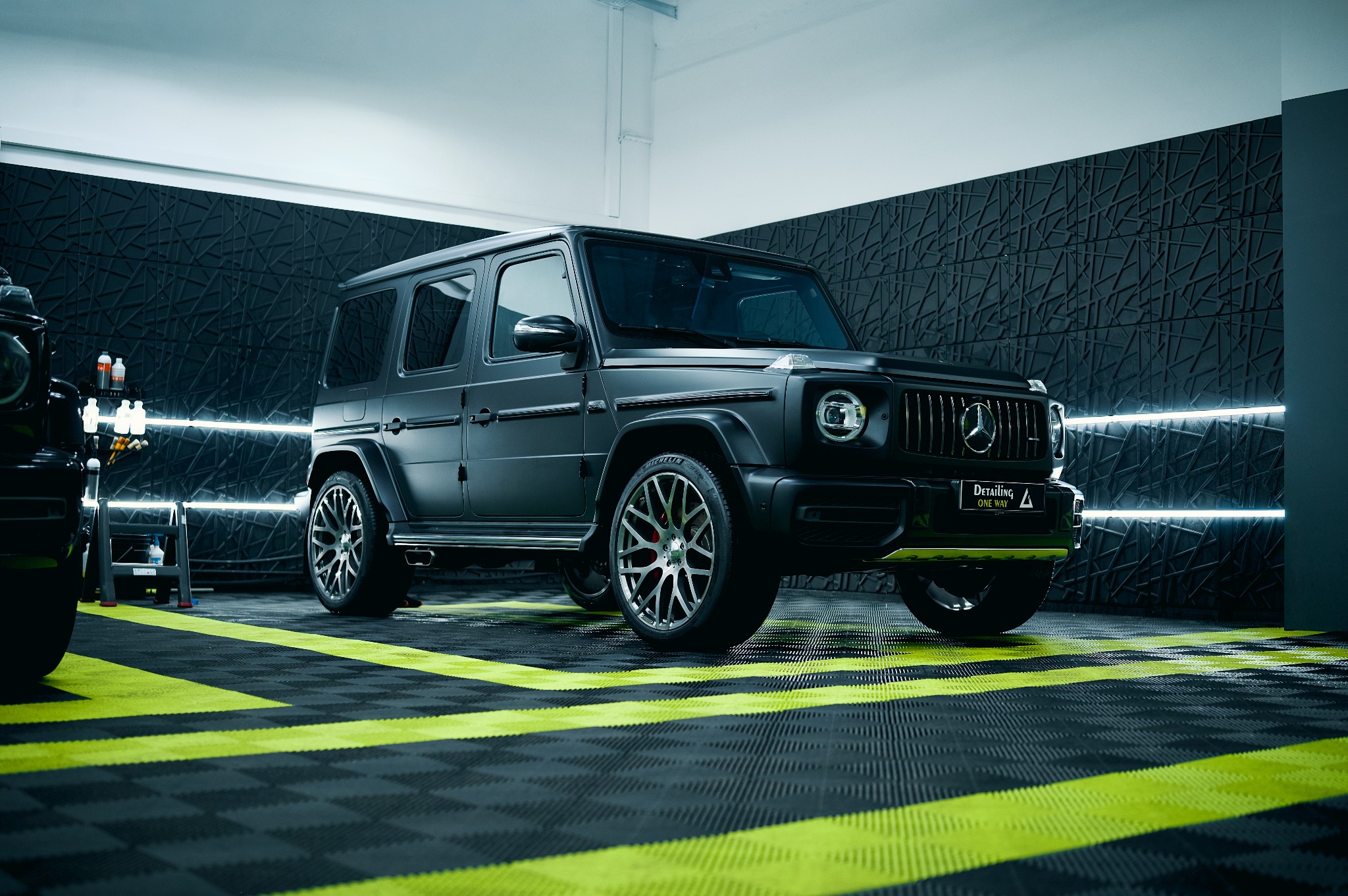 Matowy, czarny Mercedes G-klasa po detailingu w studiu. Widoczne oświetlenie LED, teksturowana ściana i podłoga w czarno-żółtą szachownicę. Profesjonalne studio detailingowe.