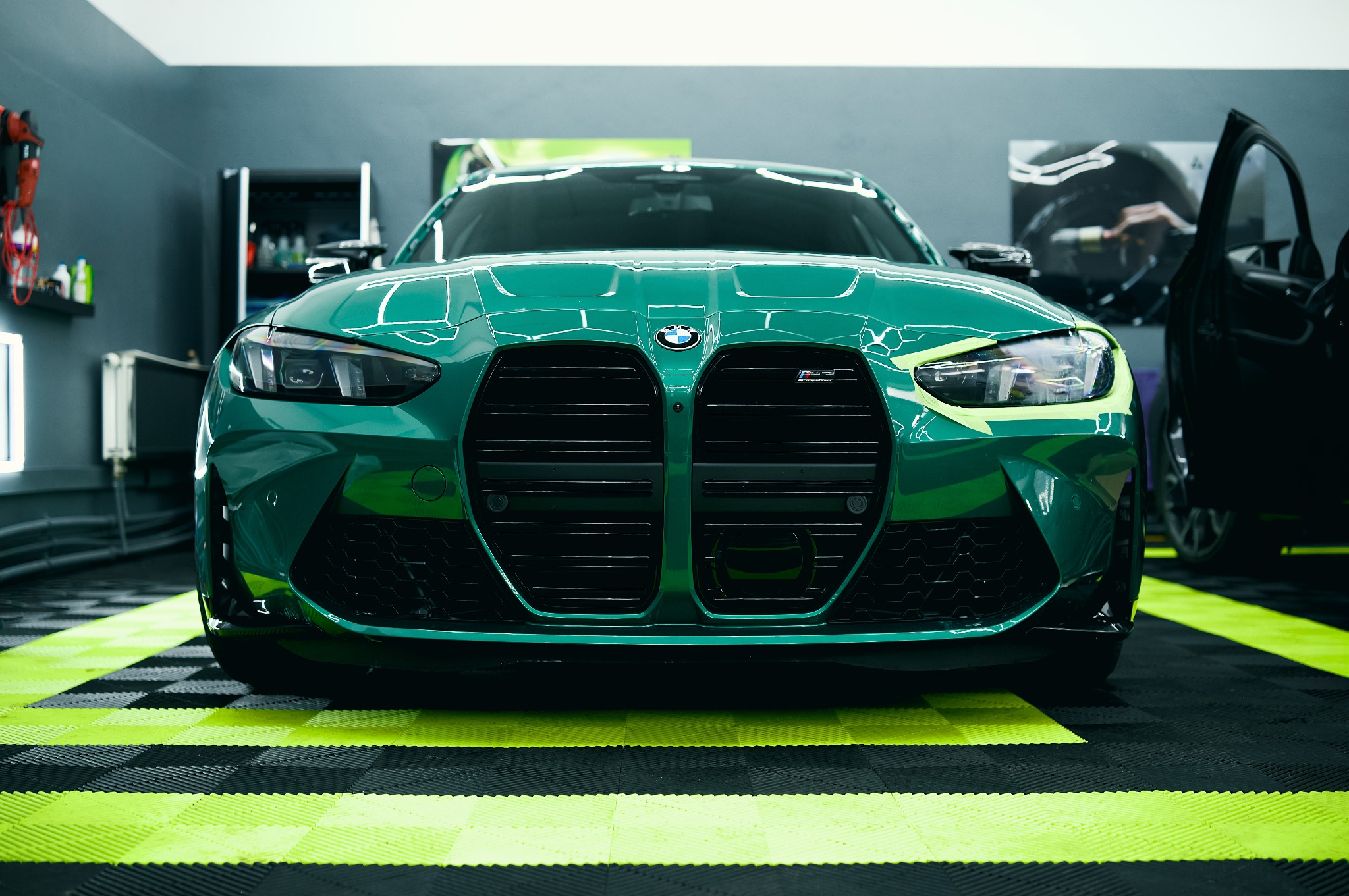 Zielony samochód BMW M4 G82 z czarnym grillem i żółtymi akcentami, przygotowywany do oklejania, widok z przodu na tle garażu z otwartymi drzwiami.