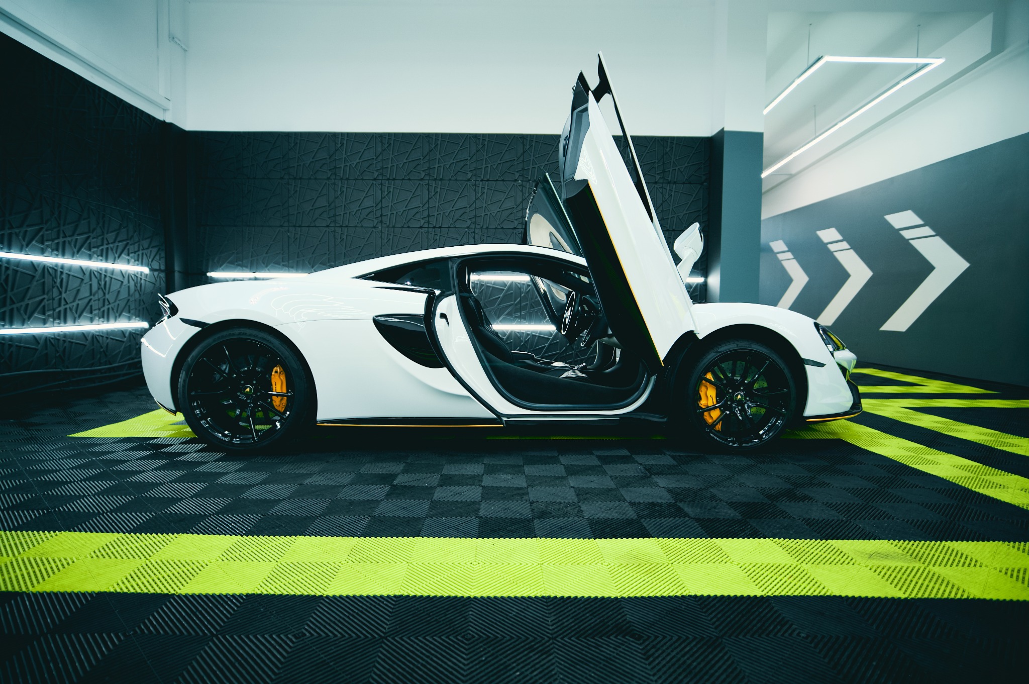 Biały sportowy samochód z otwartymi drzwiami typu 'skrzydła mewy' w nowoczesnym garażu z dekoracyjną ścianą i podłogą w czarno-zieloną szachownicę.