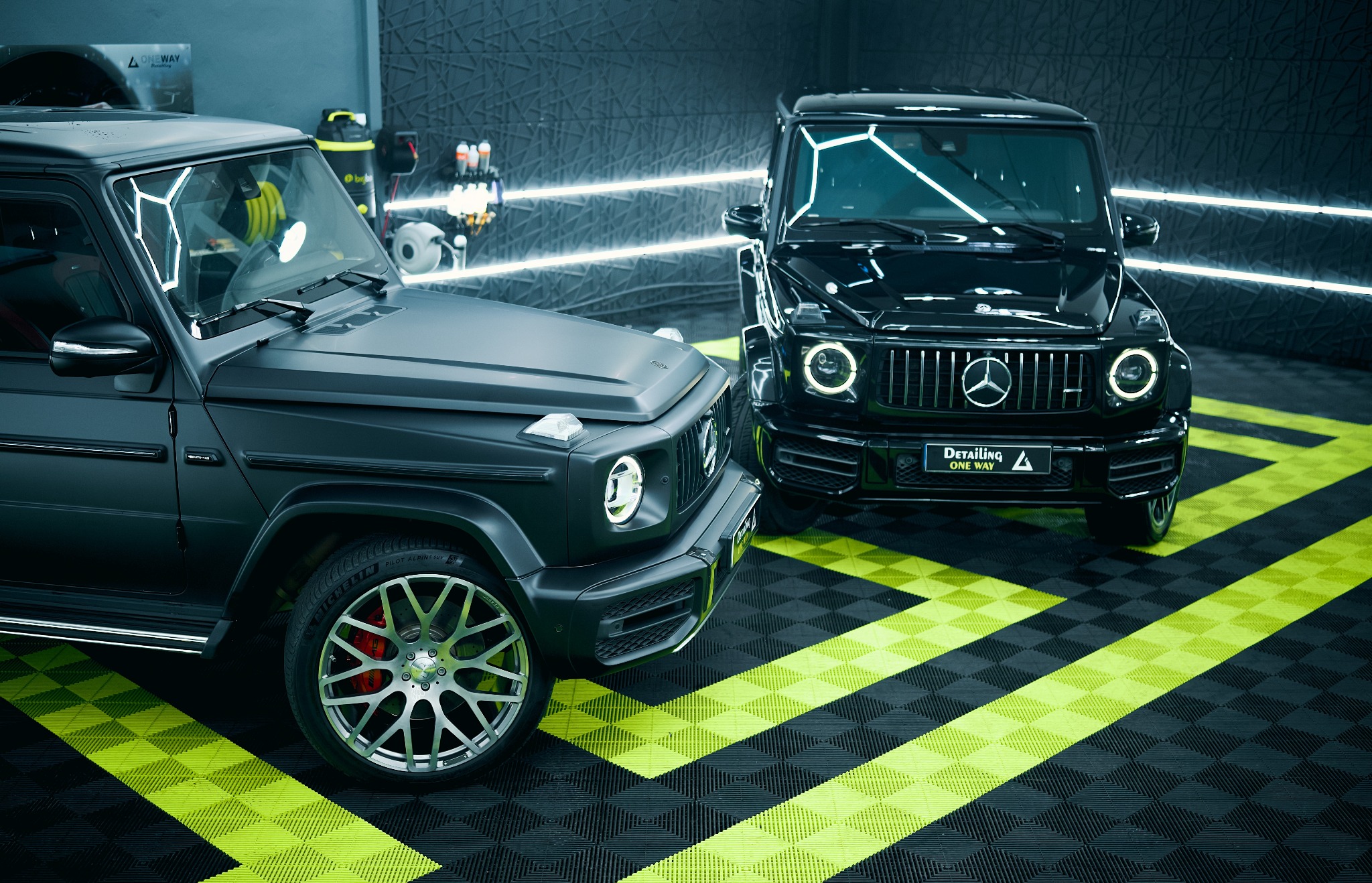 Dwa luksusowe Mercedesy klasy G: matowy szary i błyszczący czarny, stoją w studiu detailingowym z neonowym oświetleniem i podłogą w czarno-zieloną szachownicę.