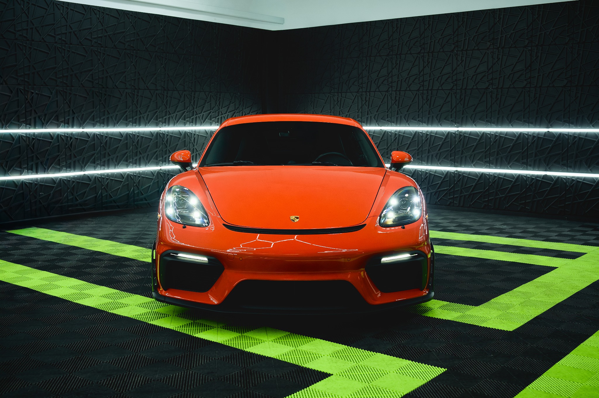 Pomarańczowe Porsche Cayman GT4 na tle czarnej ściany z neonami i zielono-czarnej podłogi. Ujęcie centralne, podkreślające sportowy charakter auta.