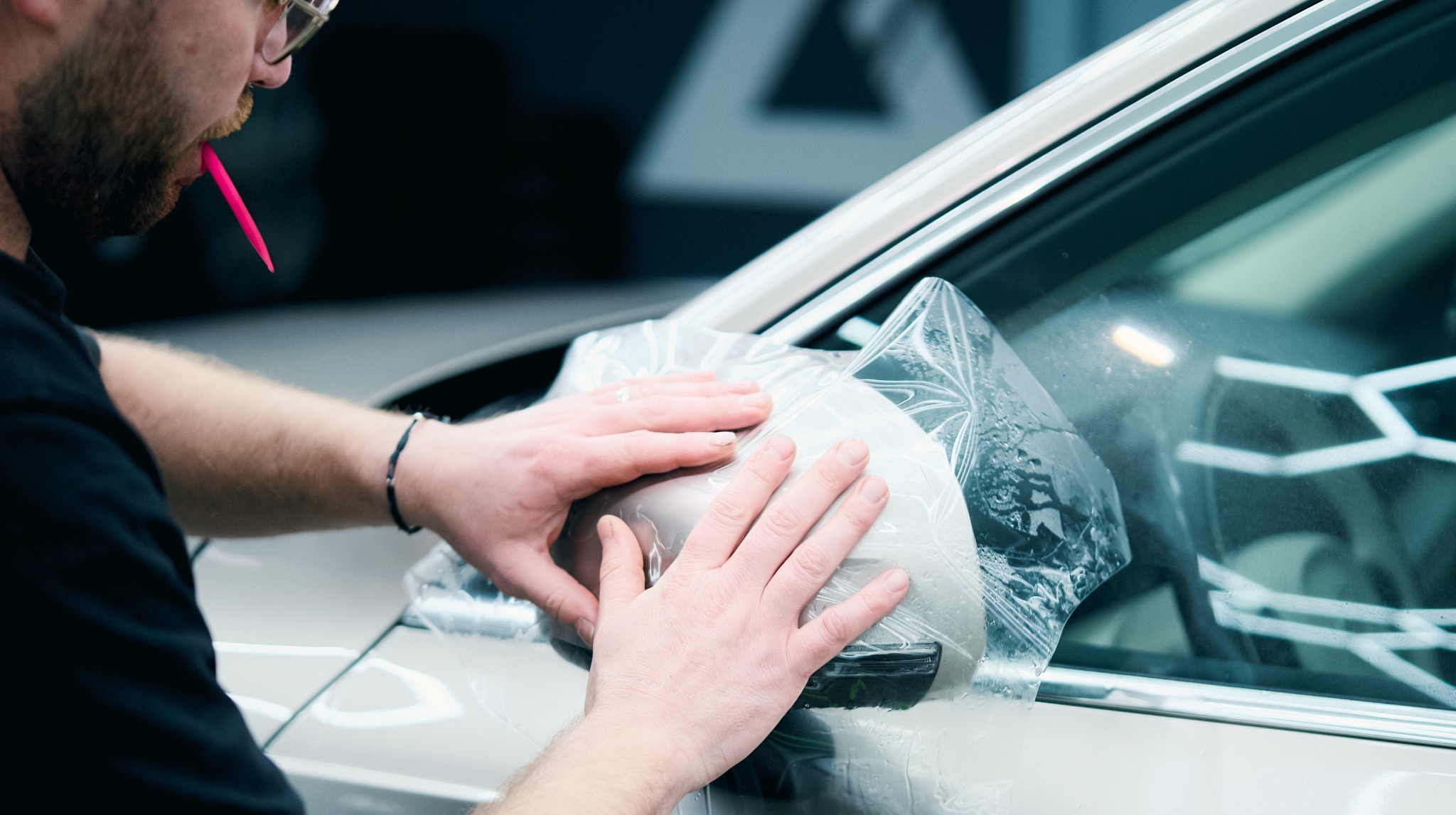 Oklejanie lusterka auta folią ochronną przez pracownika w warsztacie. Widoczne dłonie dociskające folię, fragment karoserii i narzędzie w ustach.