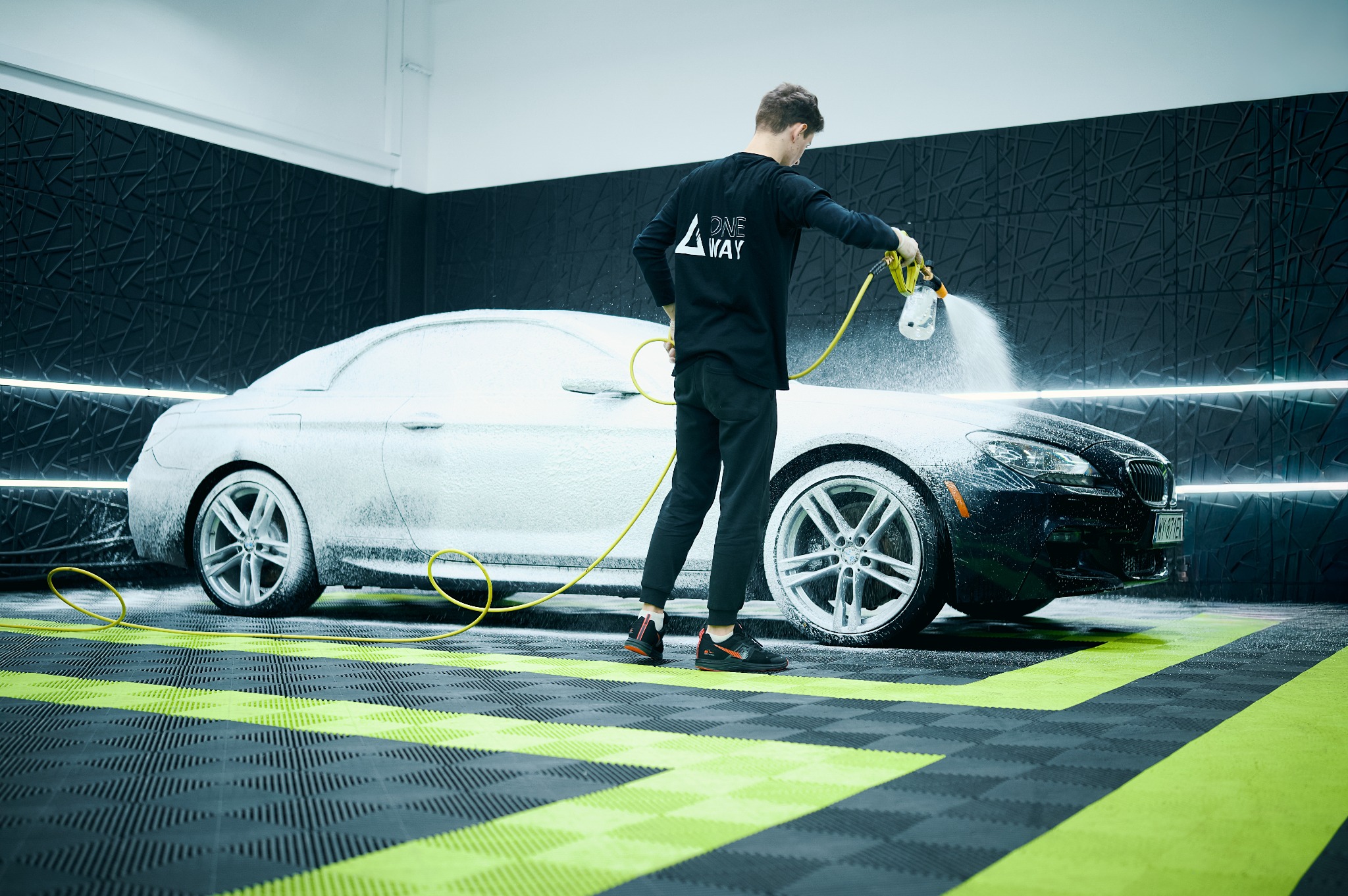 Czyszczenie pianą aktywnej na czarnym BMW serii 6 w nowoczesnej myjni z wzorzystą ścianą i podłogą. Pracownik w czarnym uniformie spryskuje auto.
