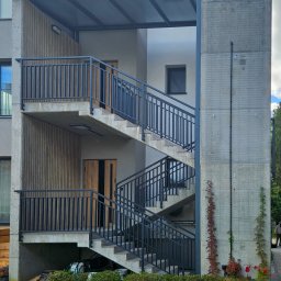 Technologia Metalu - Zewnętrzne schody betonowe z ciemnymi balustradami, prowadzące do dwóch wejść. Nad schodami zadaszenie. W tle budynek mieszkalny i zieleń.