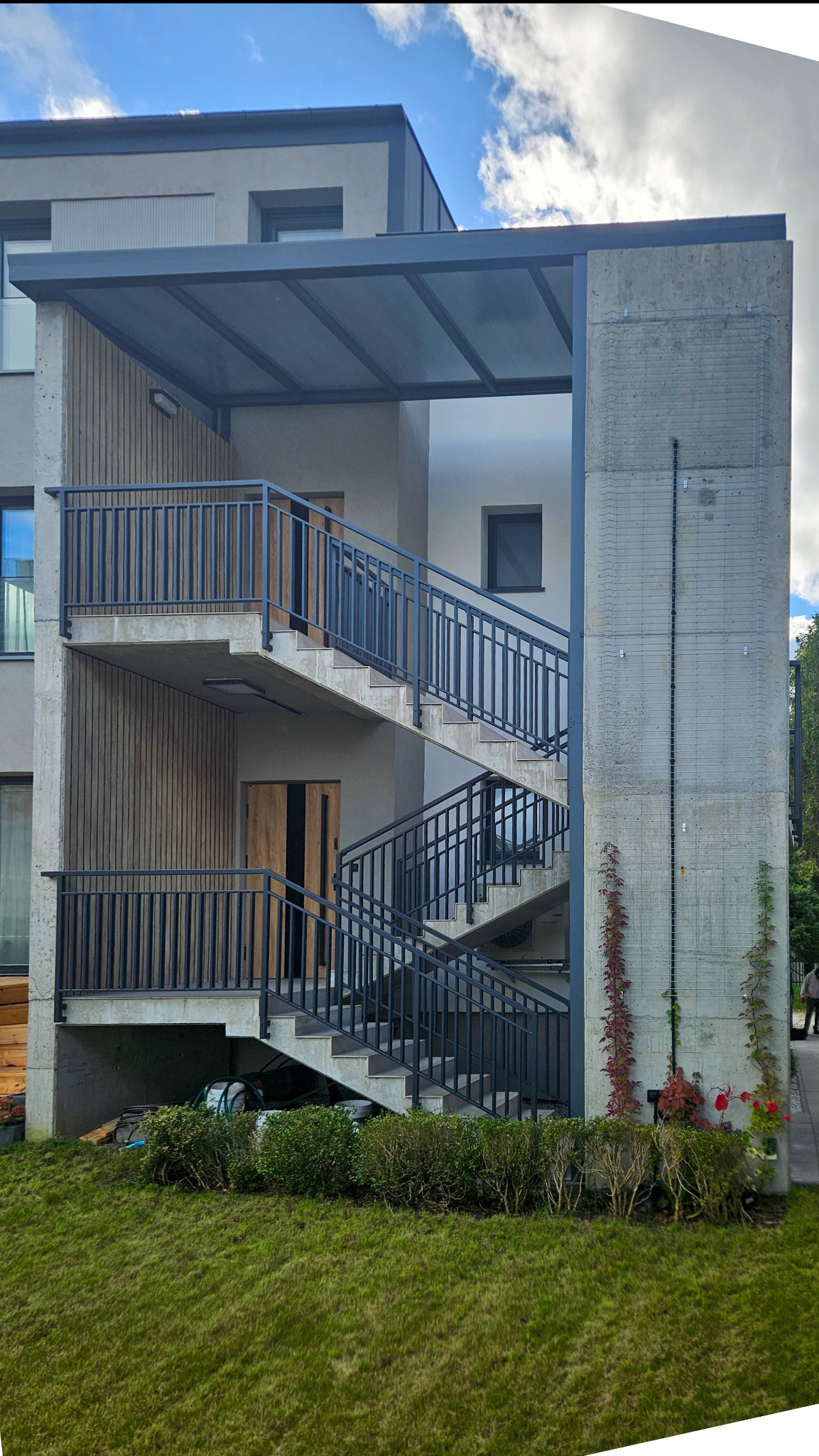 Zewnętrzne schody betonowe z ciemnymi balustradami, prowadzące do dwóch wejść. Nad schodami zadaszenie. W tle budynek mieszkalny i zieleń.