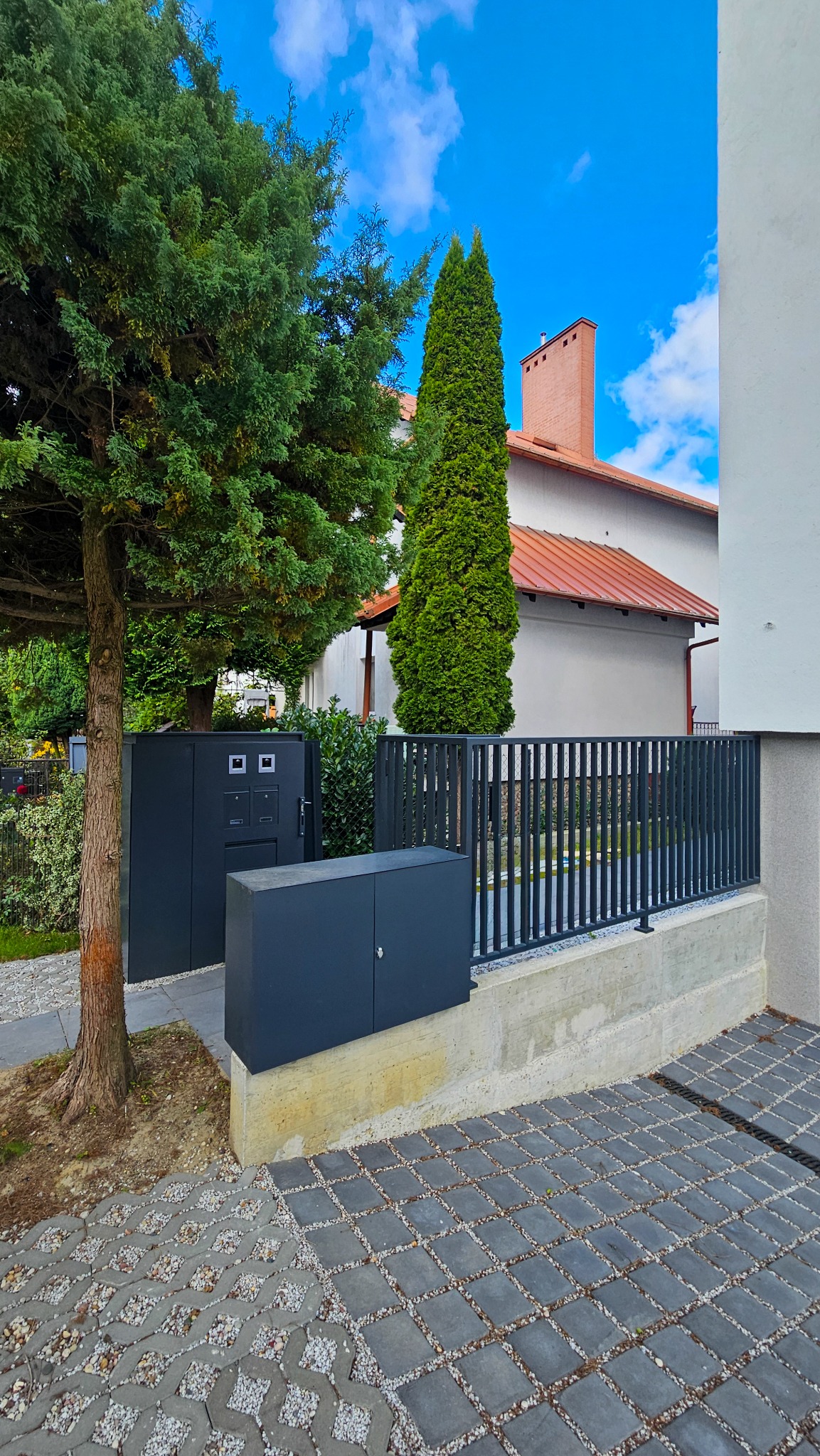 Nowoczesna, grafitowa balustrada metalowa z pionowymi szczeblami, oddzielająca posesję od chodnika. Widoczne skrzynki pocztowe i fragment domu z ceglanym kominem.