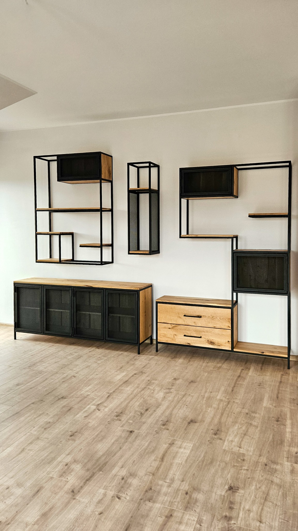 Loftowy zestaw mebli: komoda z przesuwnymi drzwiami, szafka RTV z szufladami i wiszące półki z metalowymi ramami i drewnianymi elementami na tle białej ściany.