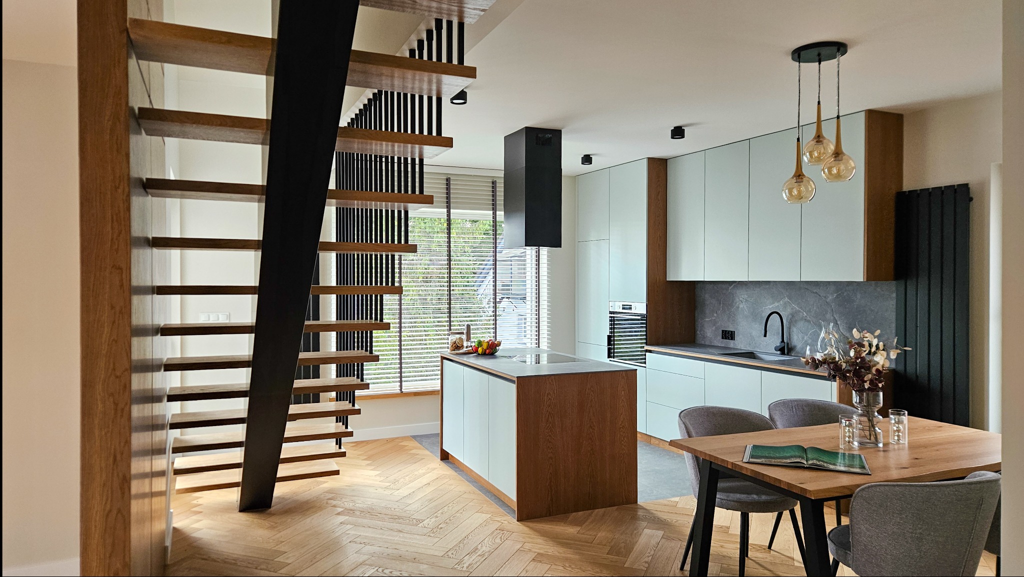 Nowoczesne schody drewniane z czarną metalową konstrukcją w jasnym wnętrzu z kuchnią i jadalnią. Parkiet w jodełkę, minimalistyczne meble kuchenne.