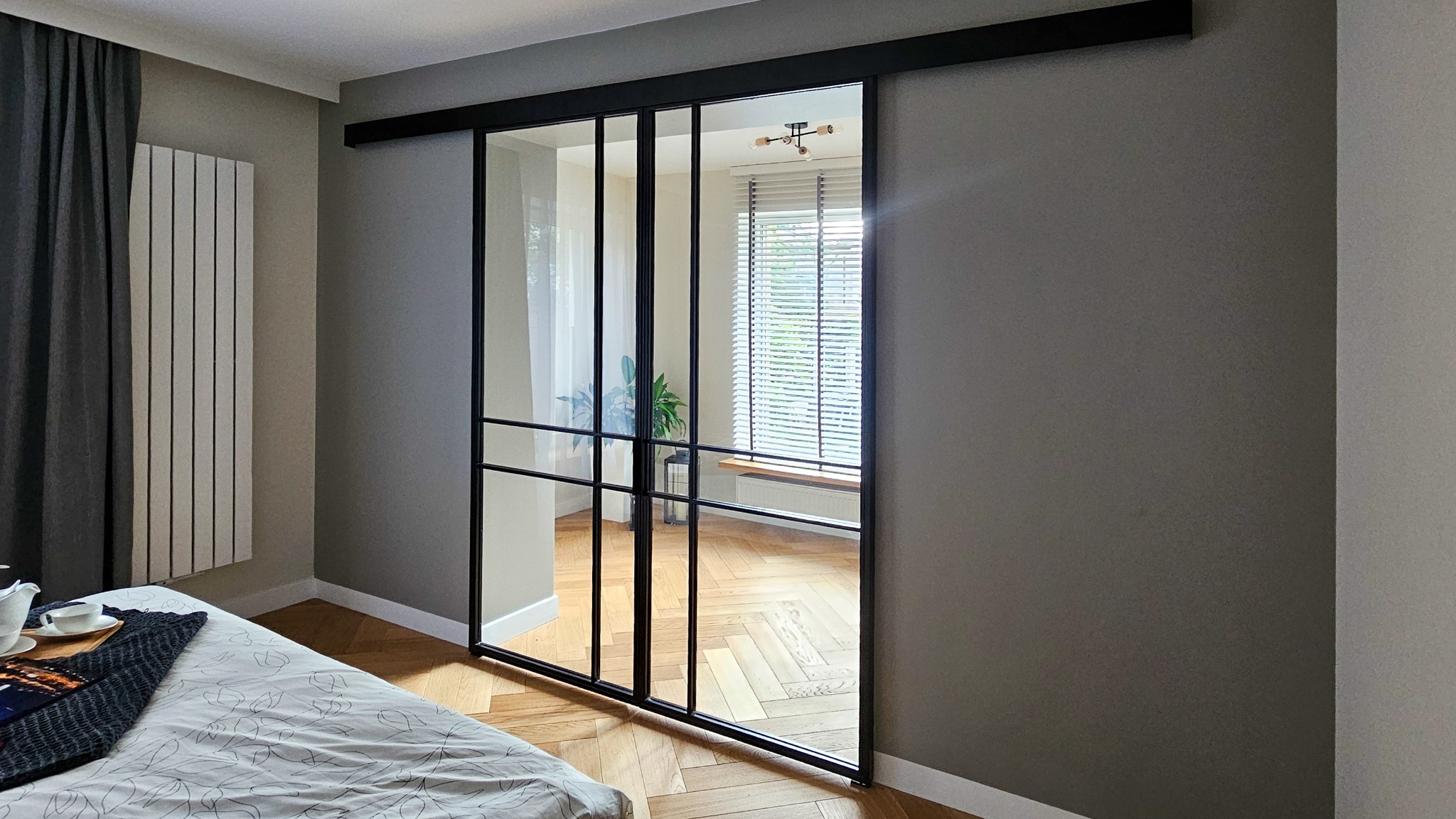 Nowoczesne, przesuwne drzwi szklane z czarną, metalową ramą, dzielone na prostokątne segmenty, widoczne z perspektywy sypialni z parkietem w jodełkę.