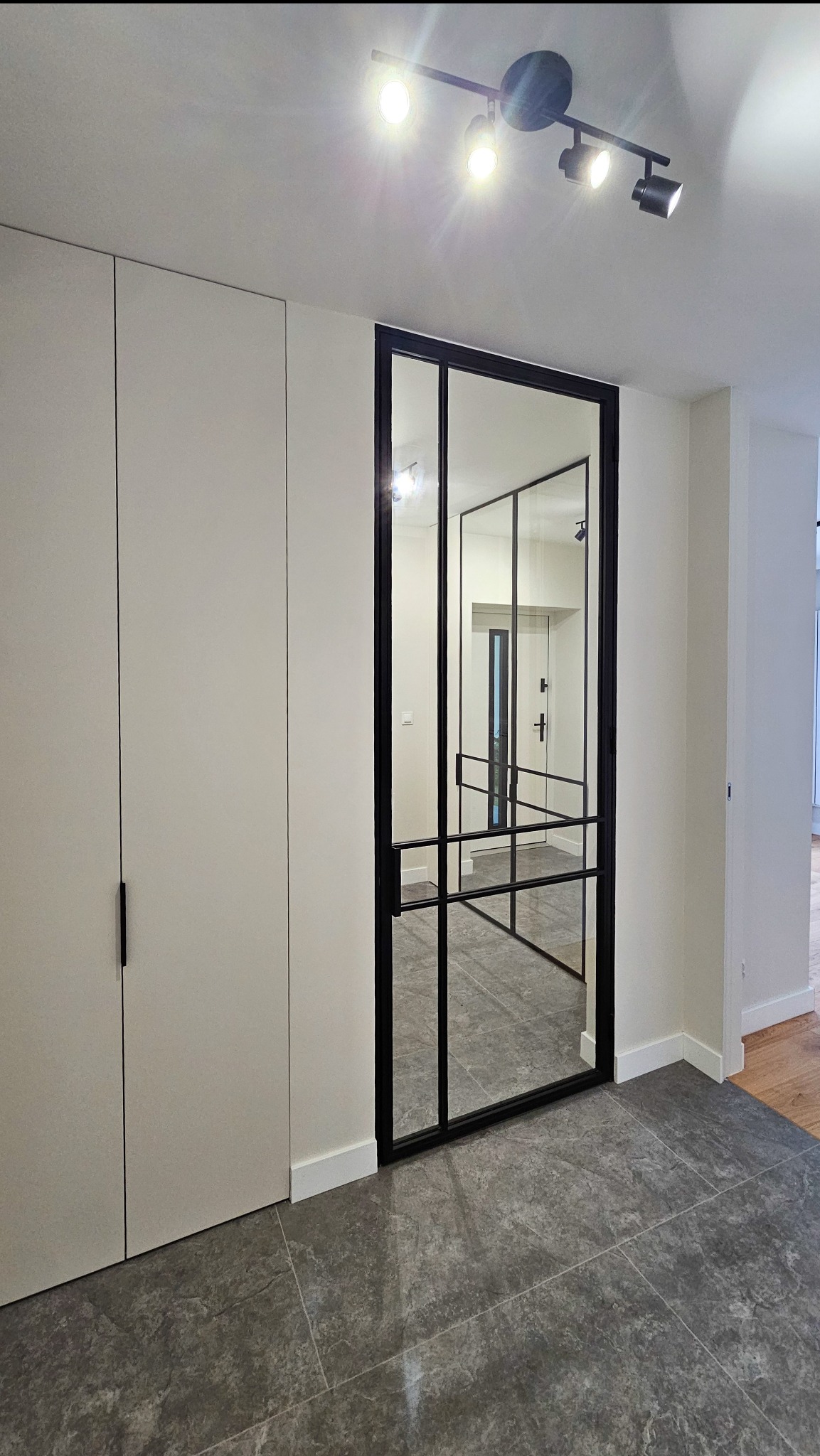 Nowoczesne, dwuskrzydłowe drzwi loftowe z czarną ramą i szklanym wypełnieniem, wkomponowane w jasne wnętrze z szafą wnękową i szarą podłogą. Widok na dalszą część mieszkania.