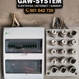 GAW-System - Tablica bezpiecznikowa z nową skrzynką elektryczną na szarej ścianie, nad nimi logo firmy Gaw-System z numerem telefonu. Widoczne wystające kable.