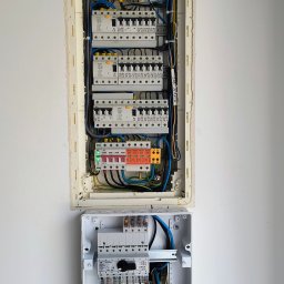 GAW-System - Dwie rozdzielnice elektryczne na białej ścianie. Górna, większa, z licznymi bezpiecznikami i okablowaniem. Dolna, mniejsza, z widocznymi przewodami i elementami zabezpieczającymi.