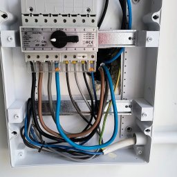 GAW-System - Otwarta, biała skrzynka rozdzielcza z widocznymi przewodami w różnych kolorach, bezpiecznikami i przełącznikiem w pozycji 'ON'. Widok z góry na instalację elektryczną.
