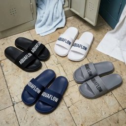 ID STUDIO - Klapki Aquaflow w czterech kolorach (czarny, biały, granatowy, szary) leżące na kafelkach, obok szafki i ręczników. Prezentacja obuwia z nadrukiem.