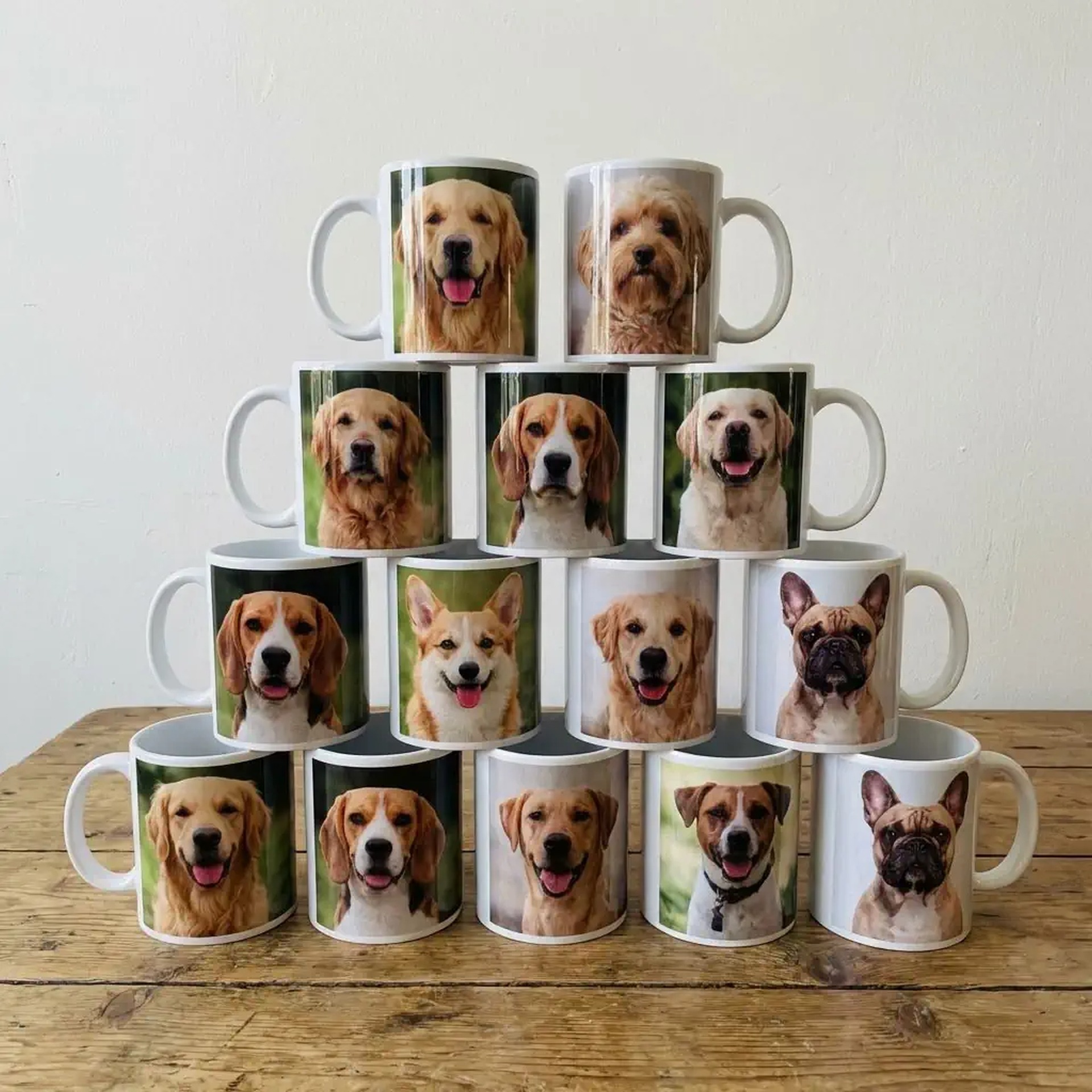 Piramida z białych kubków z nadrukami portretów psów różnych ras, ustawiona na drewnianym stole na tle białej ściany. Prezent z pupilem.