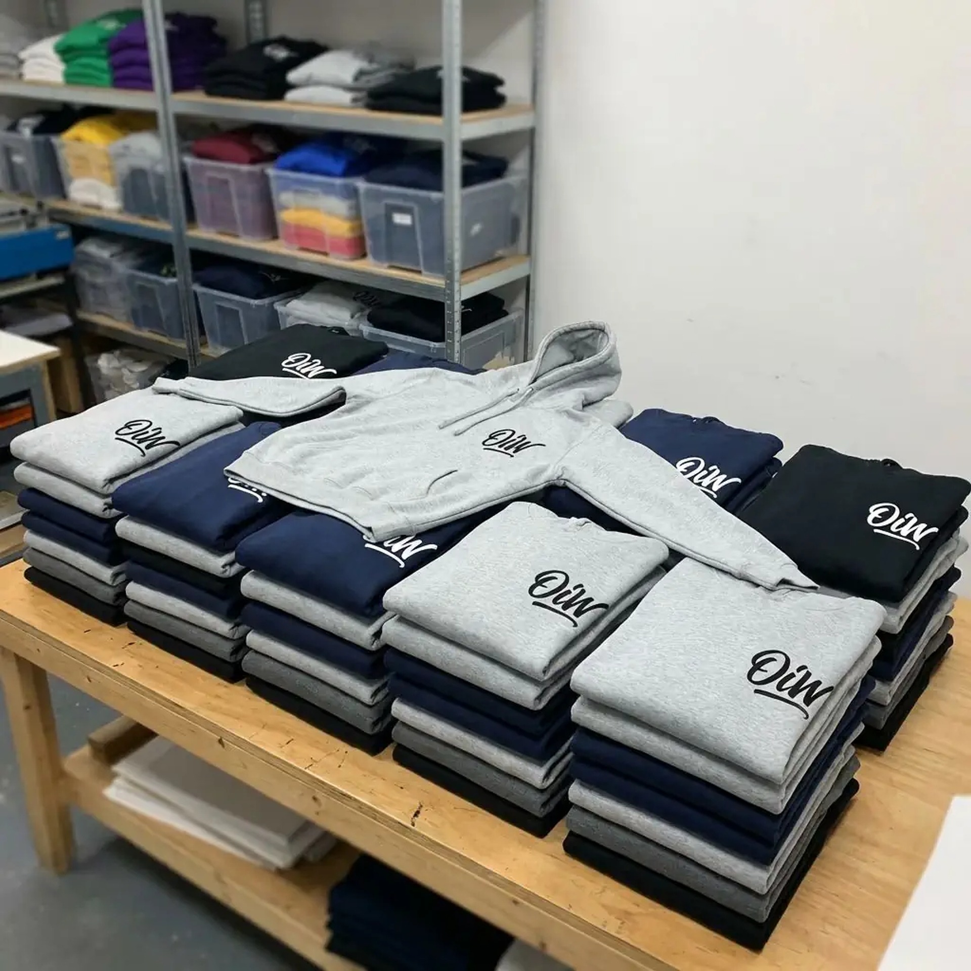 Stosy poskładanych bluz i koszulek z nadrukiem 'Oiw' w odcieniach szarości, granatu i czerni, ułożone na drewnianym stole w pracowni odzieżowej.