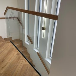 Eco Space Polska Sp. z o. o. - Szklana balustrada schodowa z drewnianym poręczą, widok z góry na parkiet w jodełkę i nowoczesne oświetlenie ścienne. Minimalistyczny design.