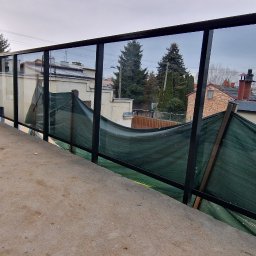 Eco Space Polska Sp. z o. o. - Szklana balustrada z czarnymi profilami na balkonie w trakcie montażu, widoczny niwelator laserowy na statywie i siatka zabezpieczająca. Tło stanowią budynki mieszkalne.