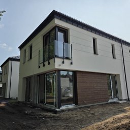 Eco Space Polska Sp. z o. o. - Nowoczesny dom z balkonem z balustradą szklaną i elewacją z drewnianymi panelami. Widoczne okna i drzwi balkonowe. Teren wokół domu w trakcie prac wykończeniowych.