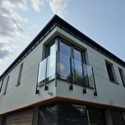 Eco Space Polska Sp. z o. o. - Nowoczesny budynek z balkonem zabezpieczonym szklaną balustradą z czarnymi elementami mocującymi, widziany z dołu na tle błękitnego nieba z chmurami.