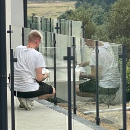 Eco Space Polska Sp. z o. o. - Montaż balustrady szklanej z czarnymi profilami na tarasie. Dwóch mężczyzn czyści szkło. W tle zieleń i elementy krajobrazu. Widok z zewnątrz.